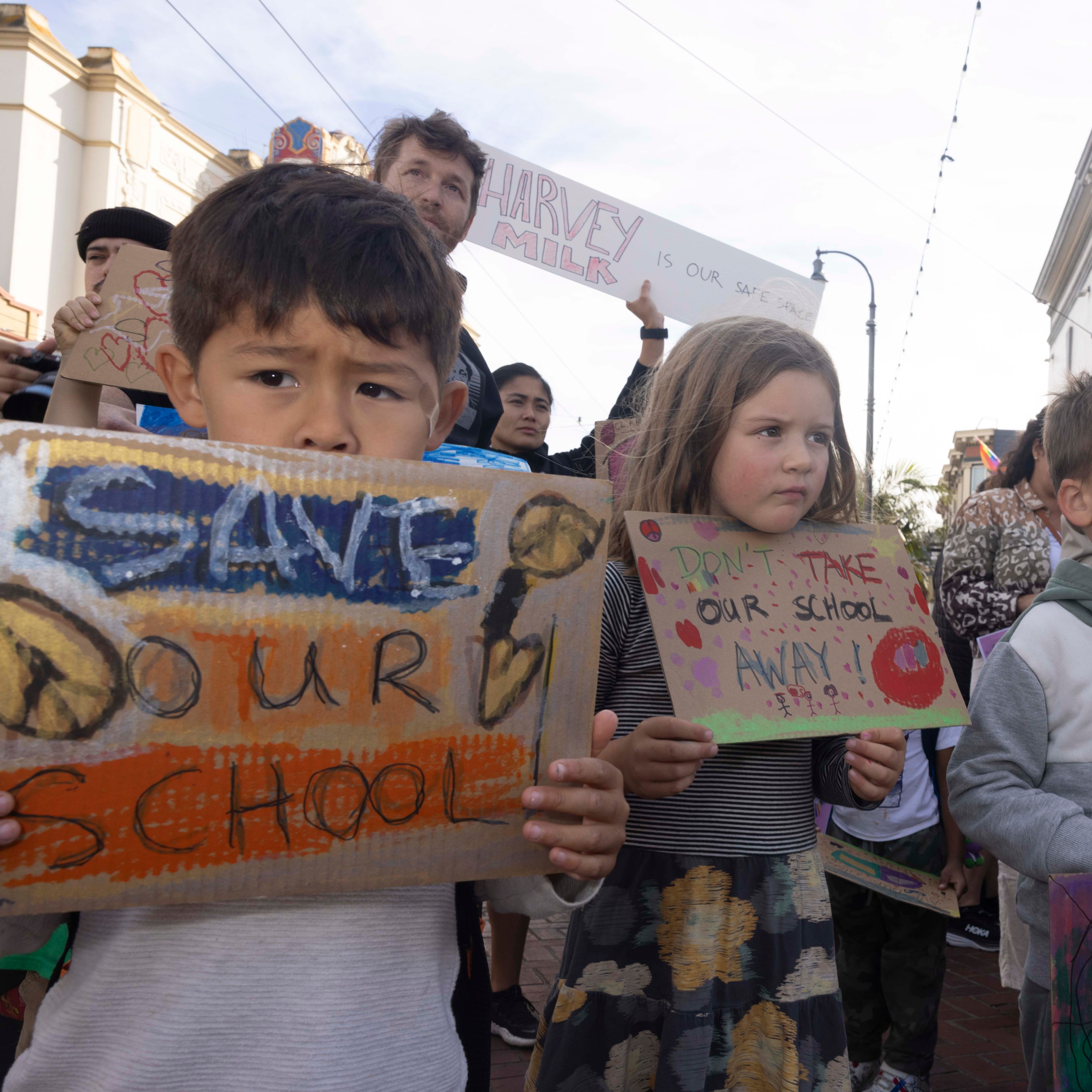 Children hold handmade signs during a protest. Their signs say “Save Our School” and “Don’t Take Our School Away!” amidst a backdrop of urban posters.