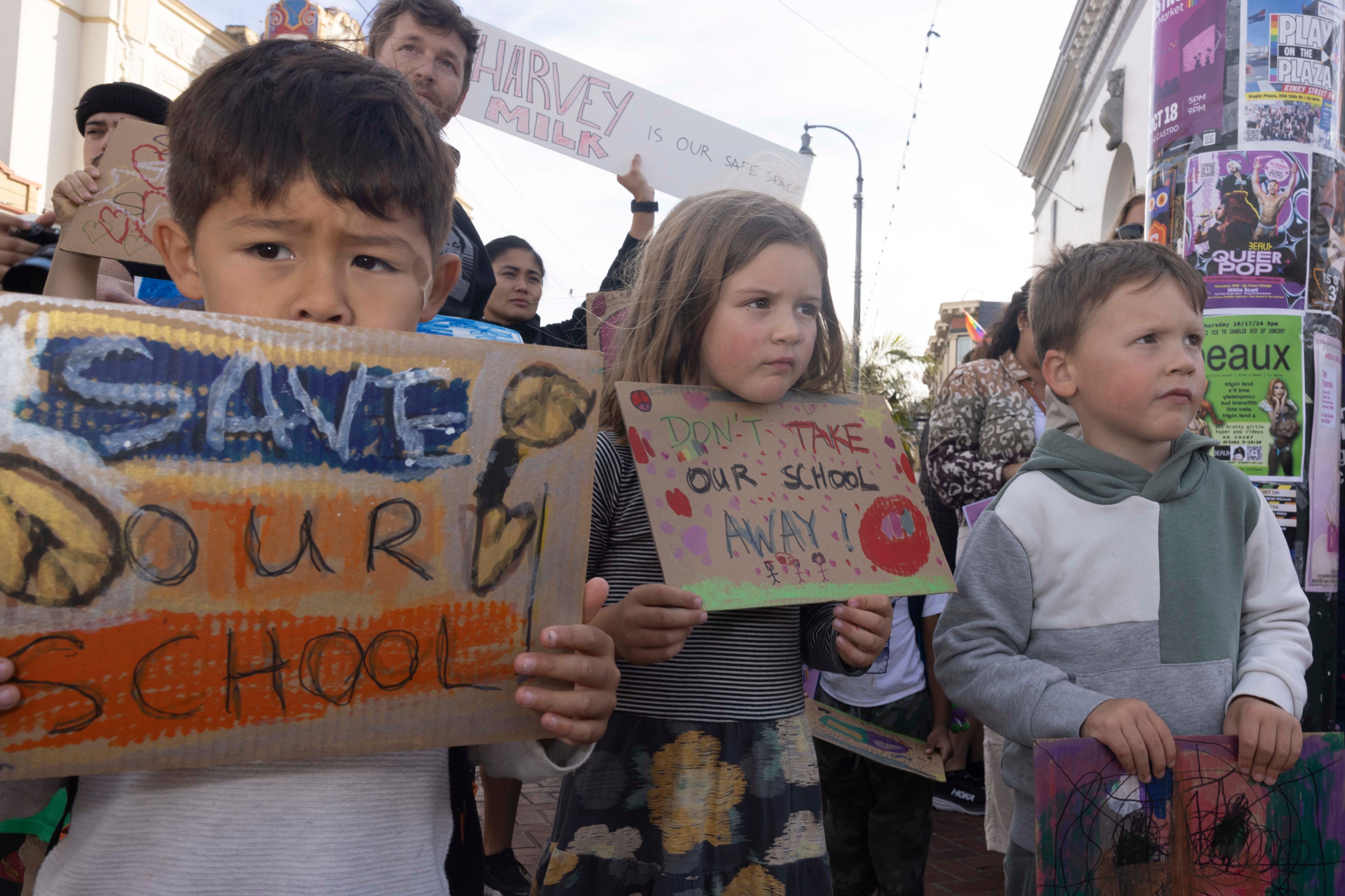 Children hold handmade signs during a protest. Their signs say “Save Our School” and “Don’t Take Our School Away!” amidst a backdrop of urban posters.