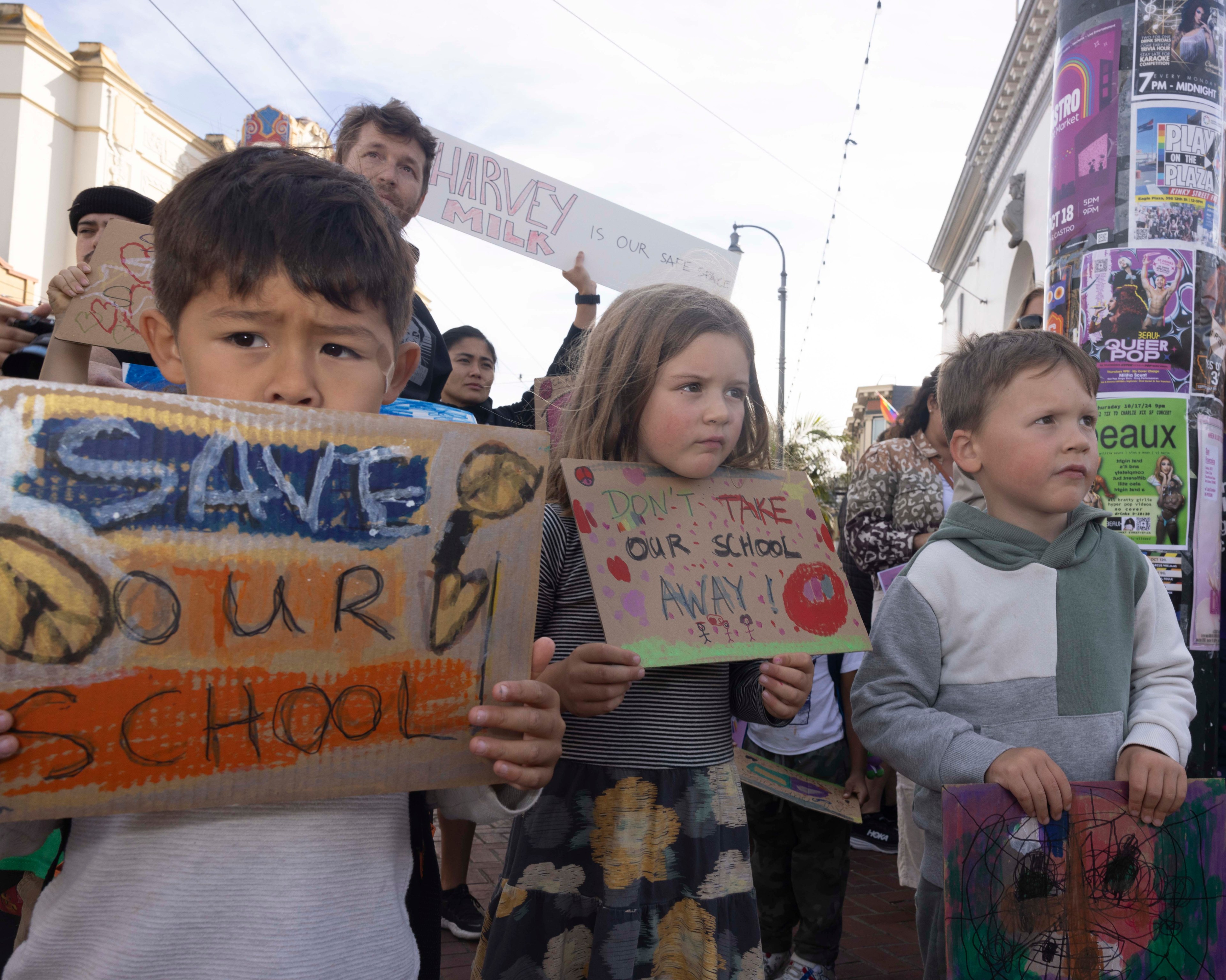 Children hold handmade signs during a protest. Their signs say “Save Our School” and “Don’t Take Our School Away!” amidst a backdrop of urban posters.