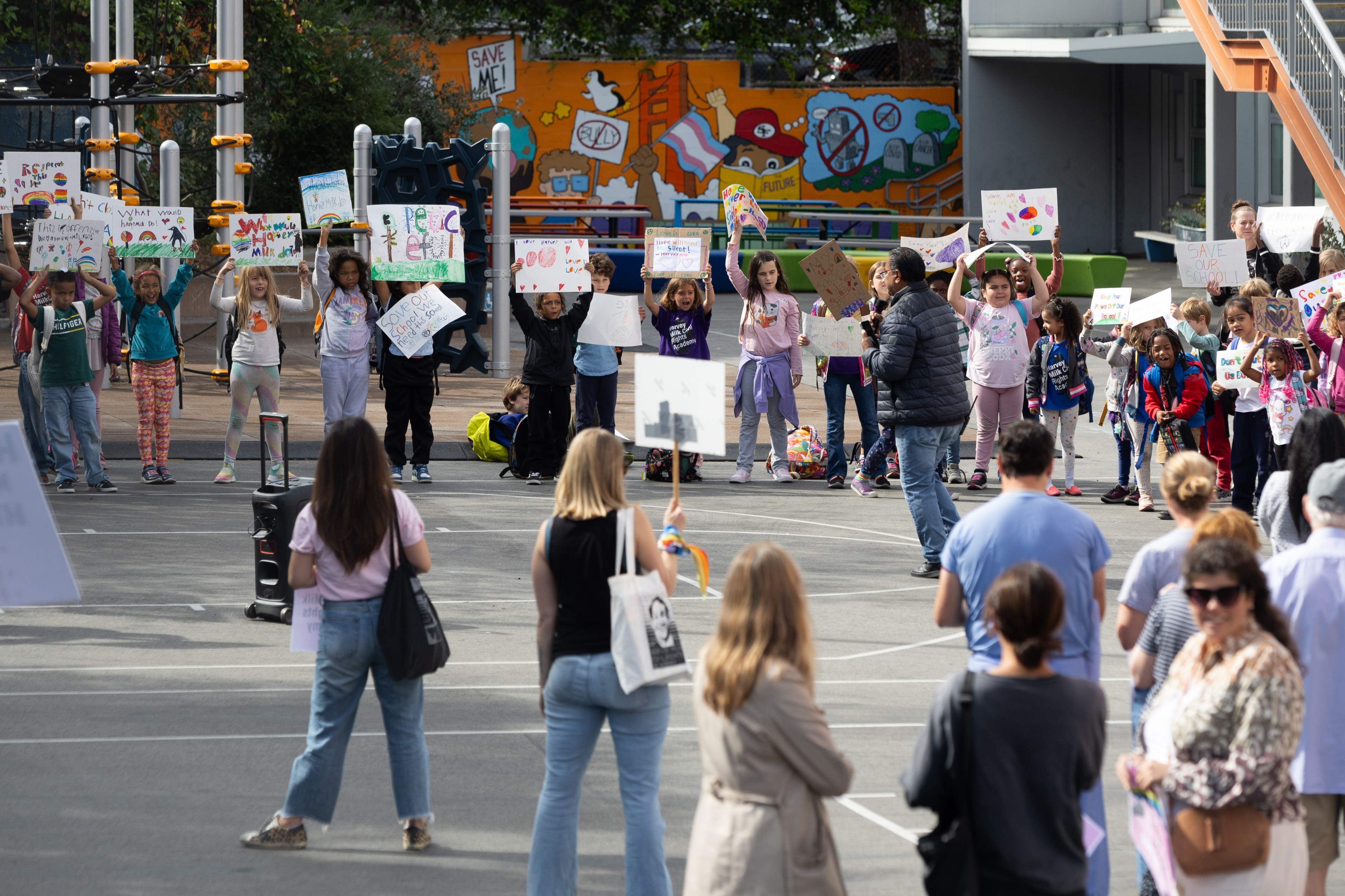 Children holding signs stand in front of a colorful mural, surrounded by adults watching them in an outdoor playground area.