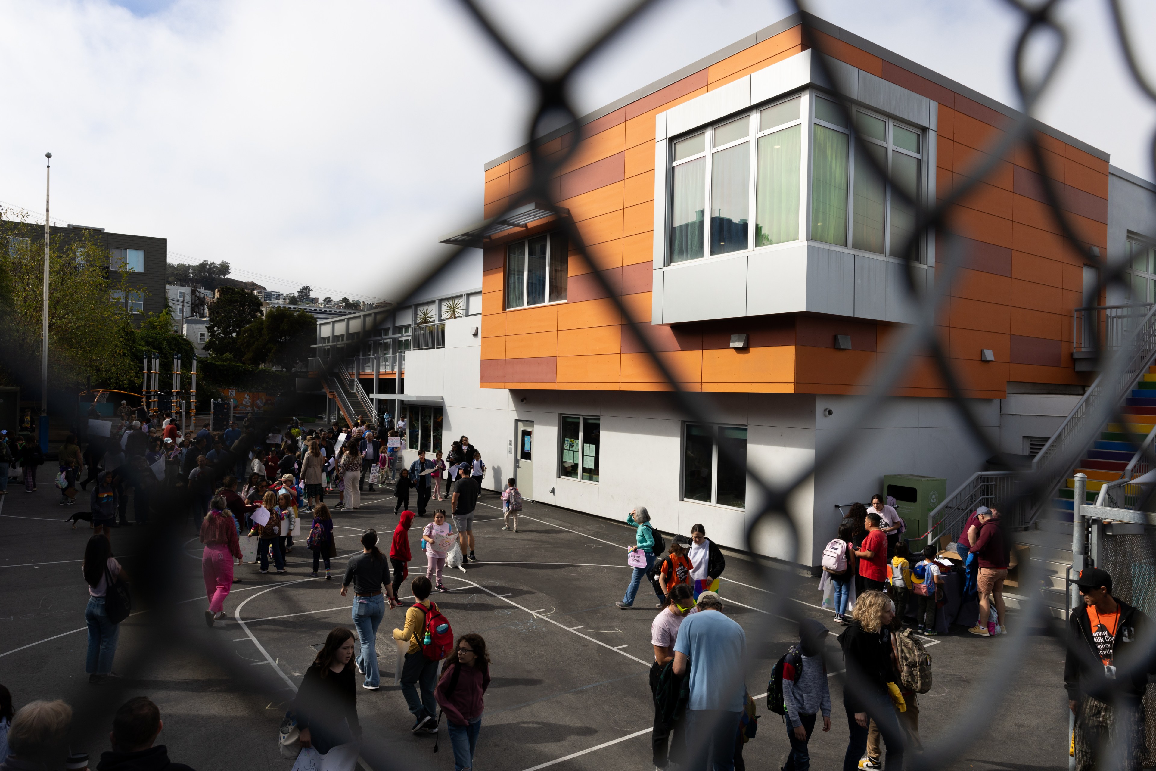 A fenced schoolyard scene shows children and adults gathered outside, with a modern orange and white building in the background. It's a casual, busy atmosphere.