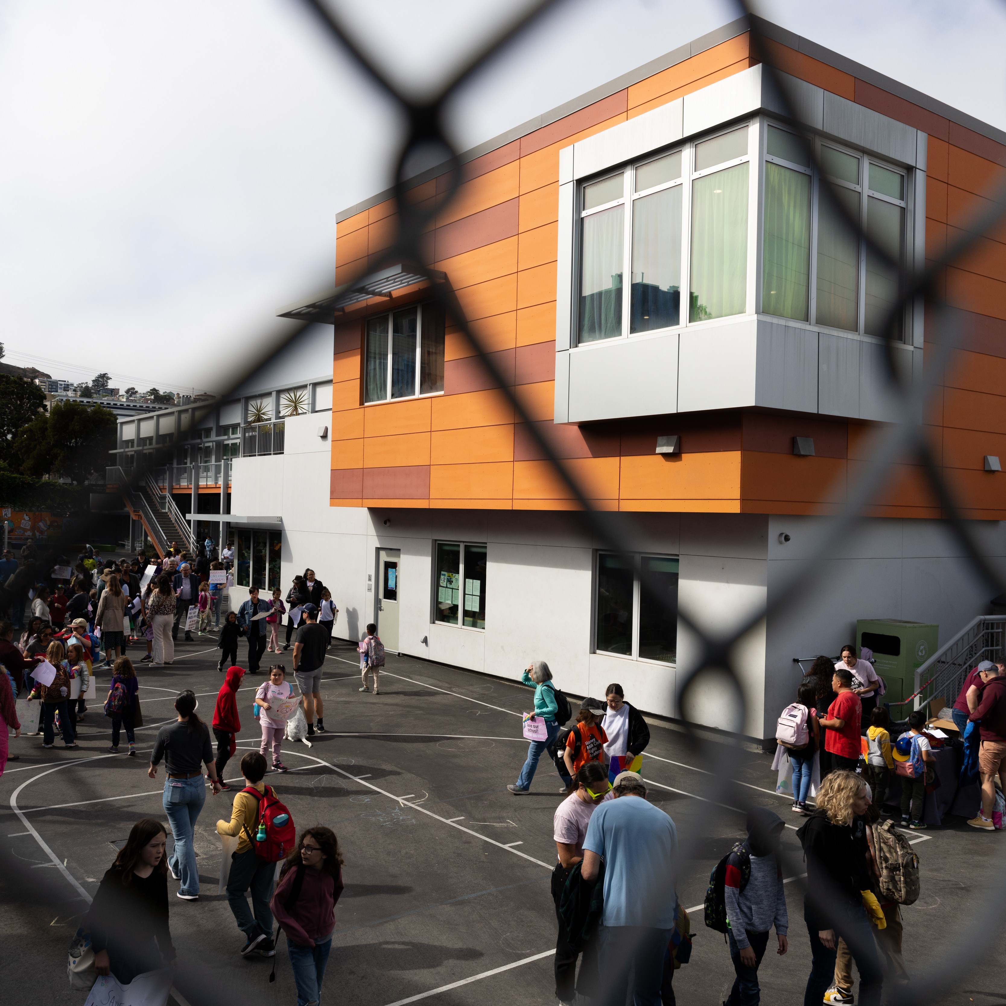A schoolyard is bustling with children and adults near a modern orange and white building. A fence partially obscures the view.