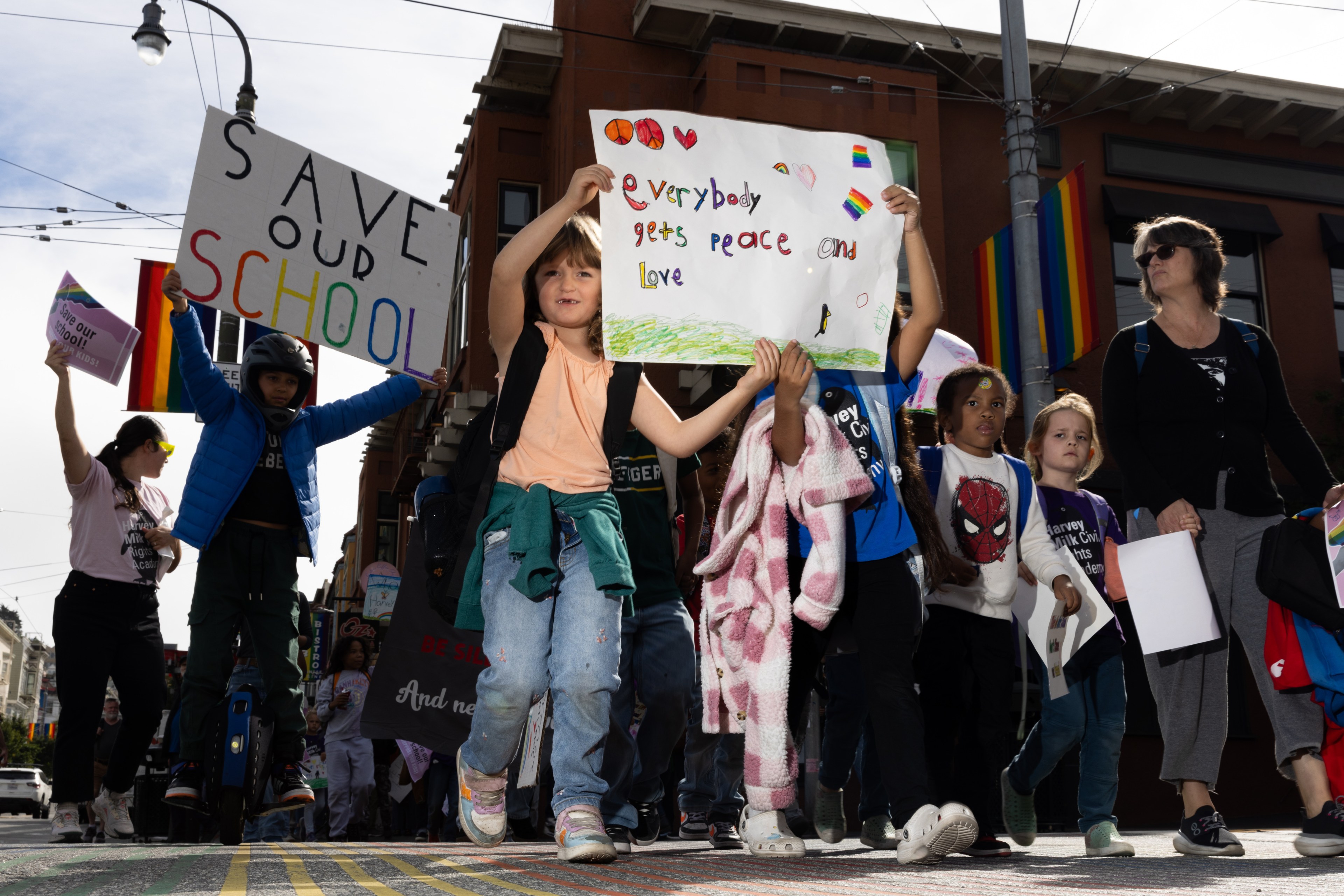 A group of people, including children, are marching with signs that say "SAVE OUR SCHOOL" and "Everybody gets peace and love," amidst rainbow flags in a city street.