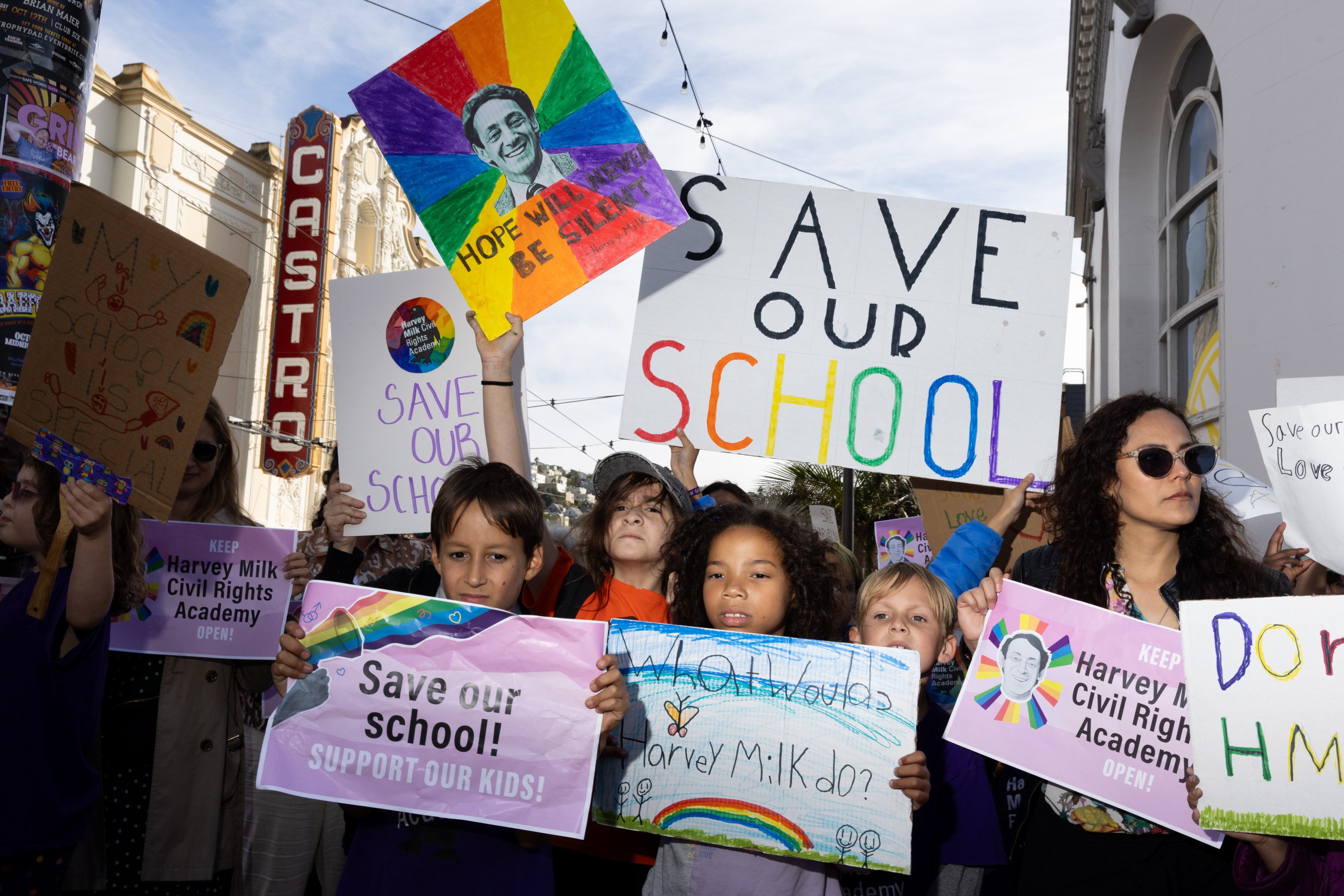 A group of people, including children, hold colorful signs advocating to &quot;Save Our School&quot; near the Castro Theatre, with rainbow themes and messages of hope.