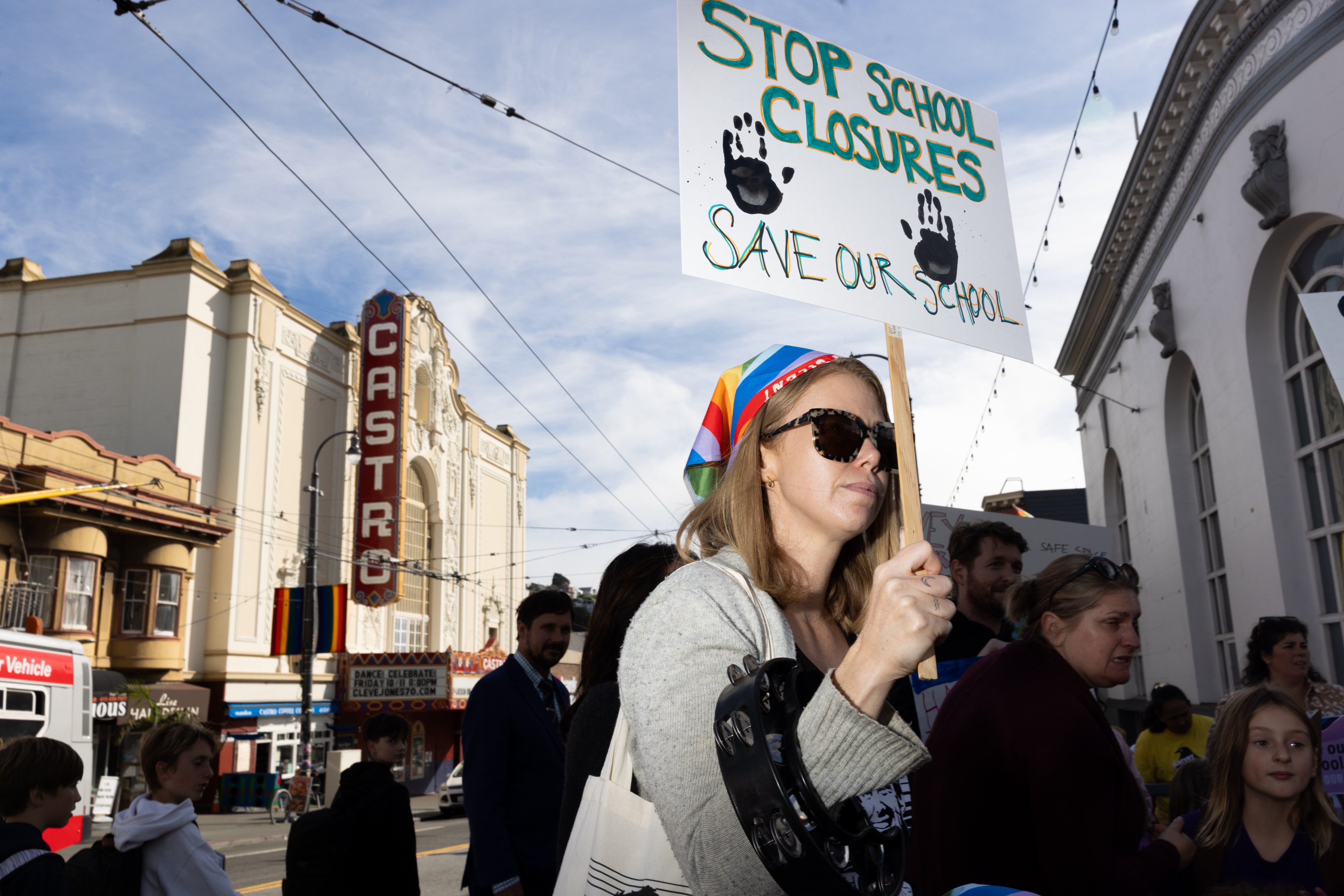 A woman holding a sign reading &quot;Stop School Closures, Save Our School&quot; stands in a crowd. The backdrop features the Castro Theater and a rainbow flag.