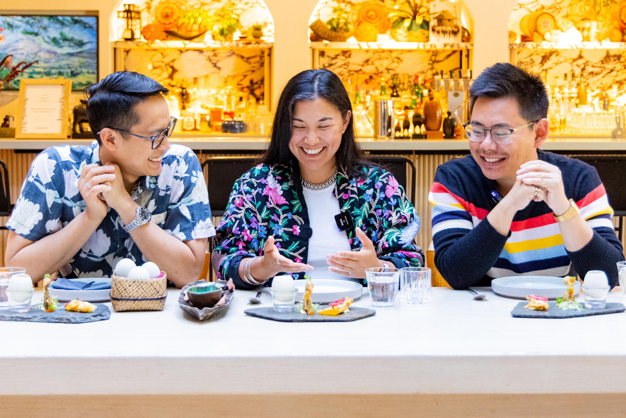 Three people are sitting at a table, smiling and conversing. The table has plates, drinks, and decorative items. The background features a warmly lit bar area.