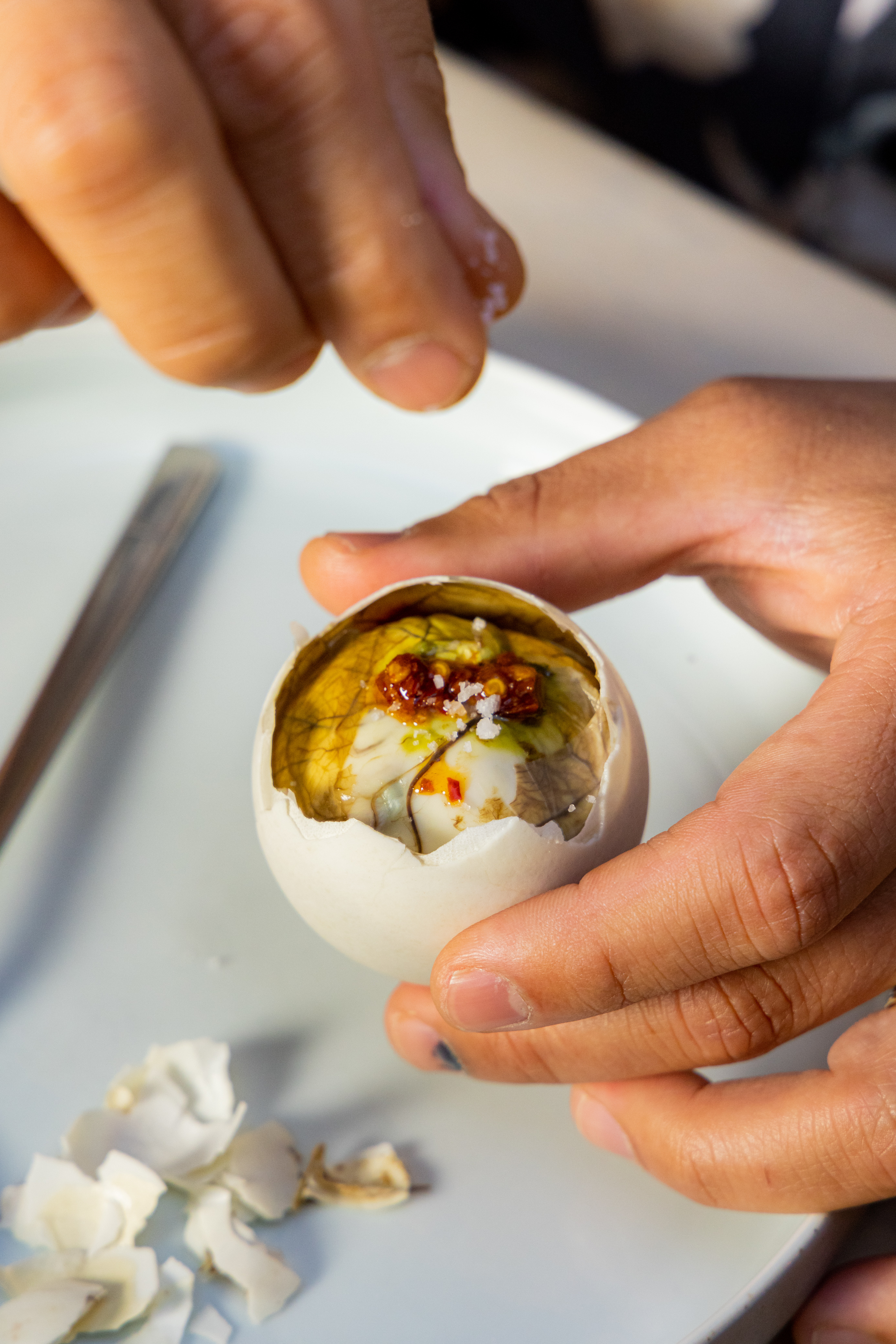 A person holds a partially peeled egg with a visible embryo inside, seasoned with salt and spices, over a plate with shell fragments.