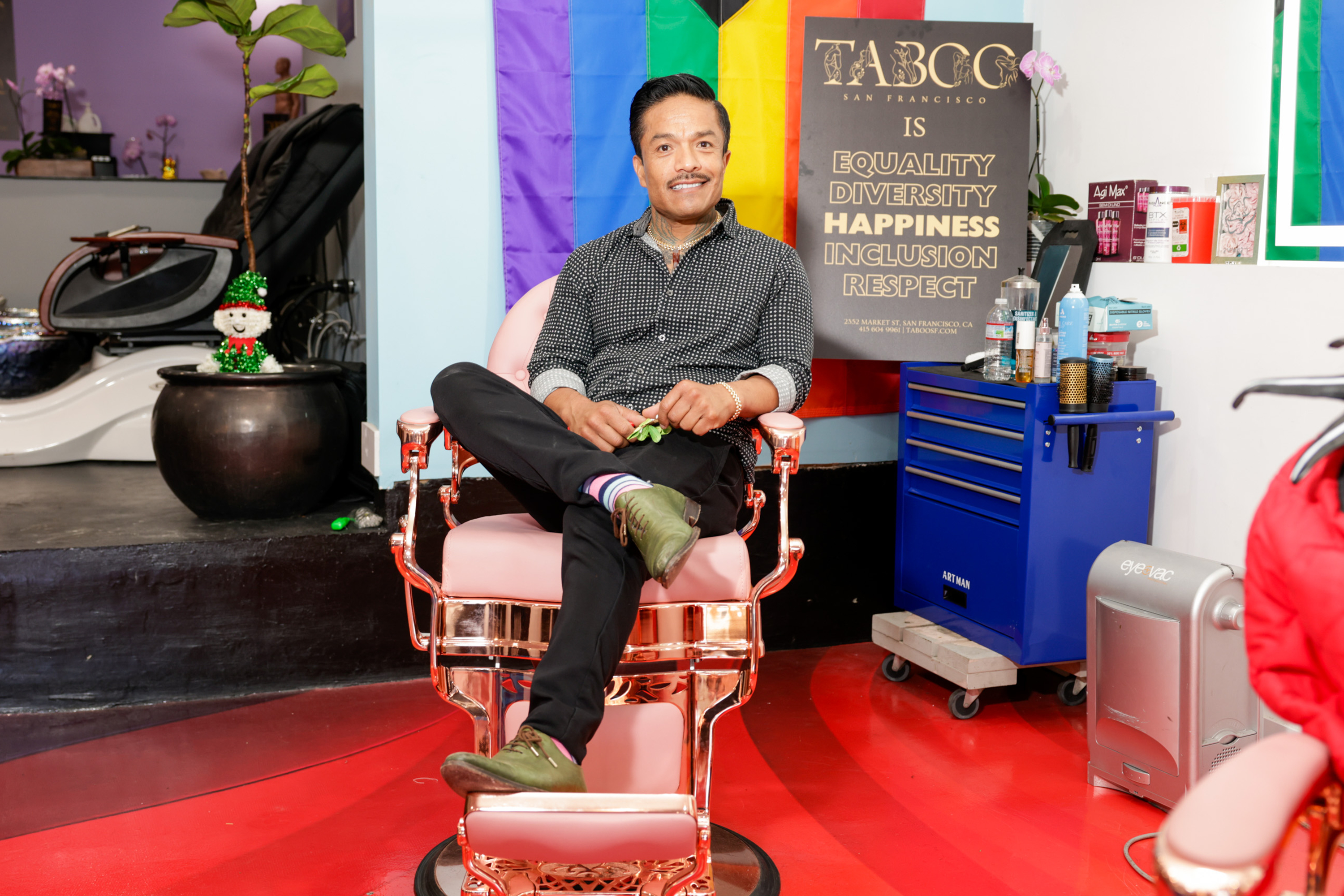 A person sits cross-legged in a barber chair, smiling. Behind them is a sign promoting equality and diversity, and a colorful flag. The environment is bright and welcoming.