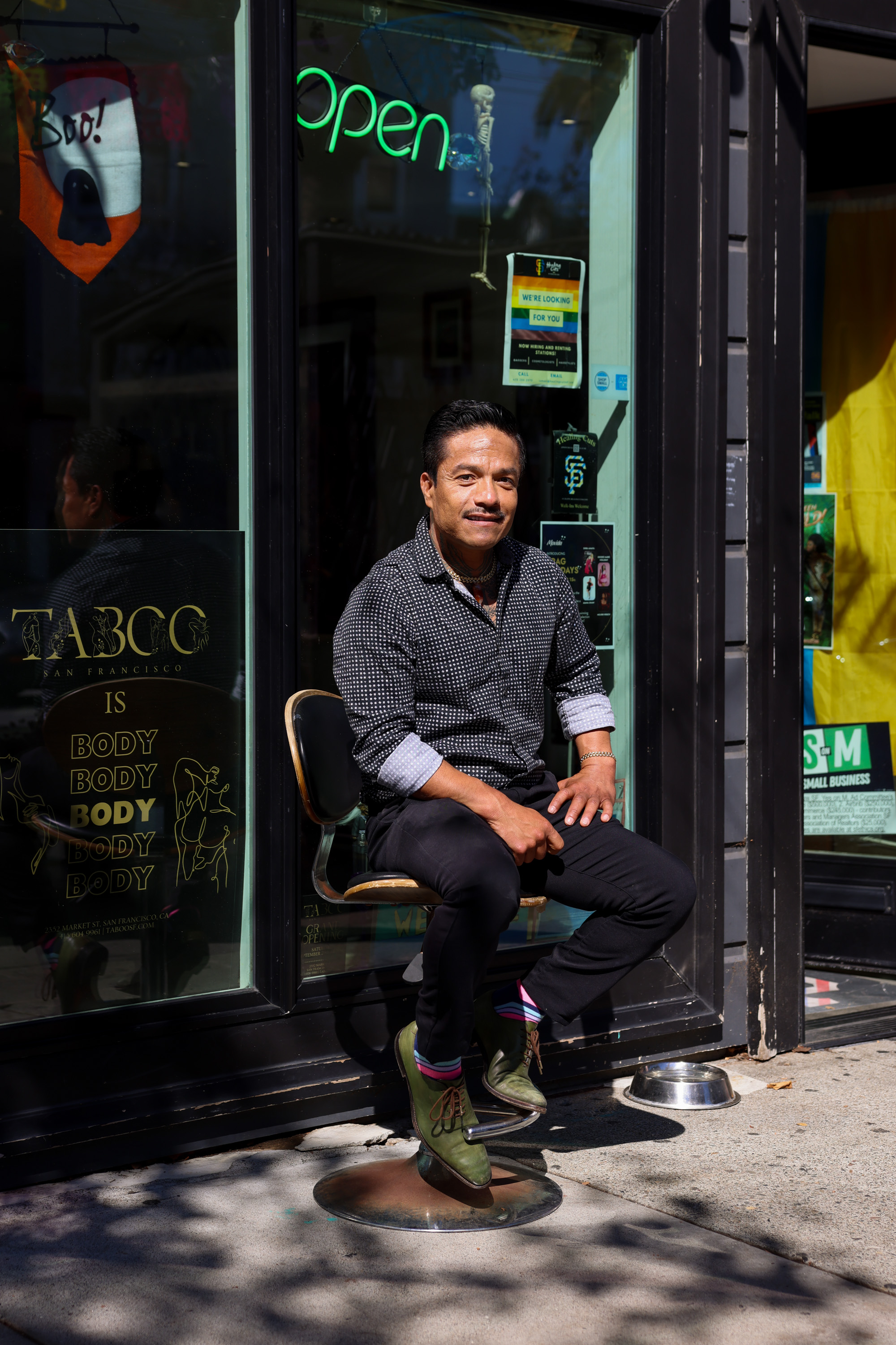 A man sits on a stool outside a storefront with a neon &quot;open&quot; sign, wearing a patterned shirt and green shoes. A &quot;Boo!&quot; sign hangs nearby.