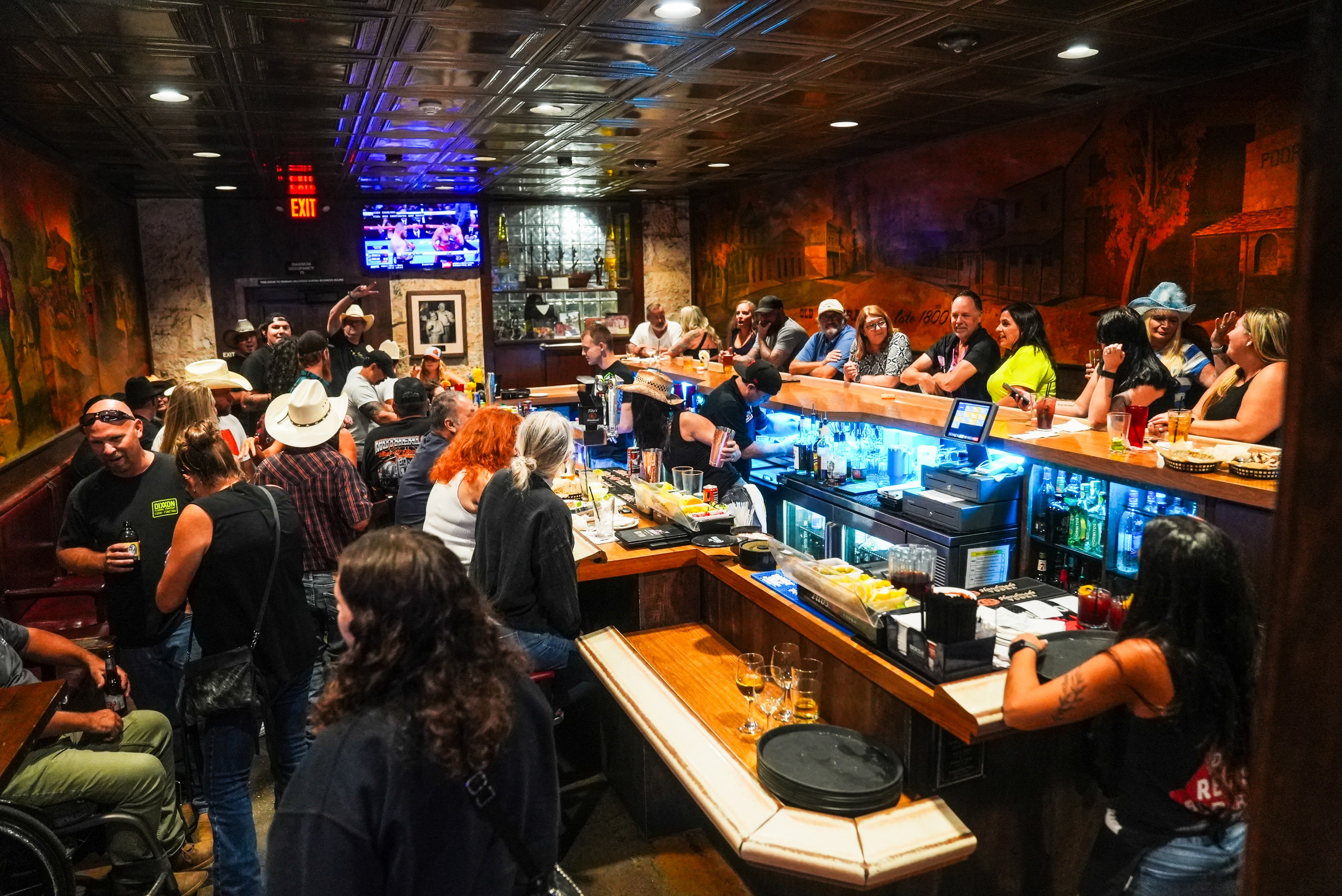 A lively bar scene with people socializing around a U-shaped counter. Several wear cowboy hats. A TV is on, and drinks are visible on the bar top.