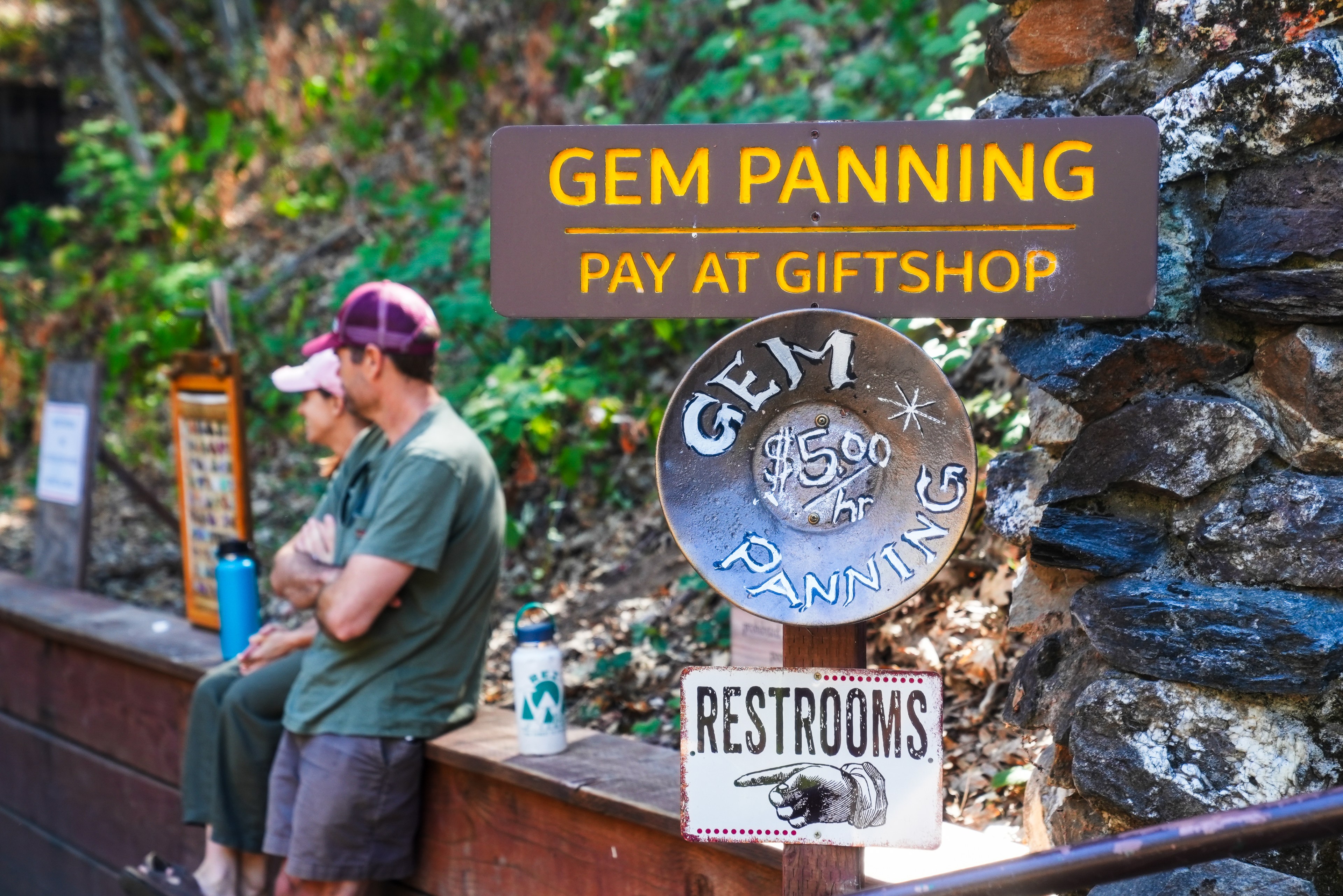 A sign for gem panning reads &quot;Pay at Gift Shop&quot; and &quot;$5.00/hr,&quot; with another sign indicating restrooms. Two people sit nearby, surrounded by greenery.