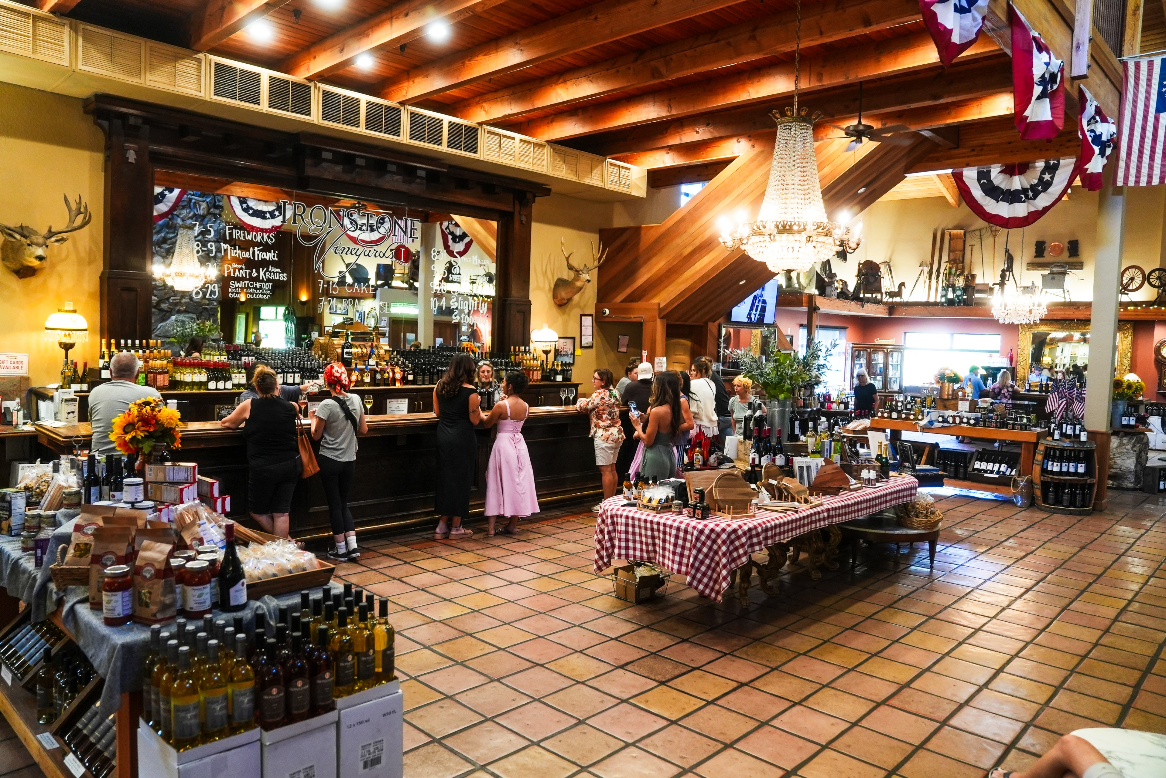 The image shows a cozy wine tasting room with a wooden bar, patrons enjoying wine, and tables displaying bottles and packaged goods under warm lighting.