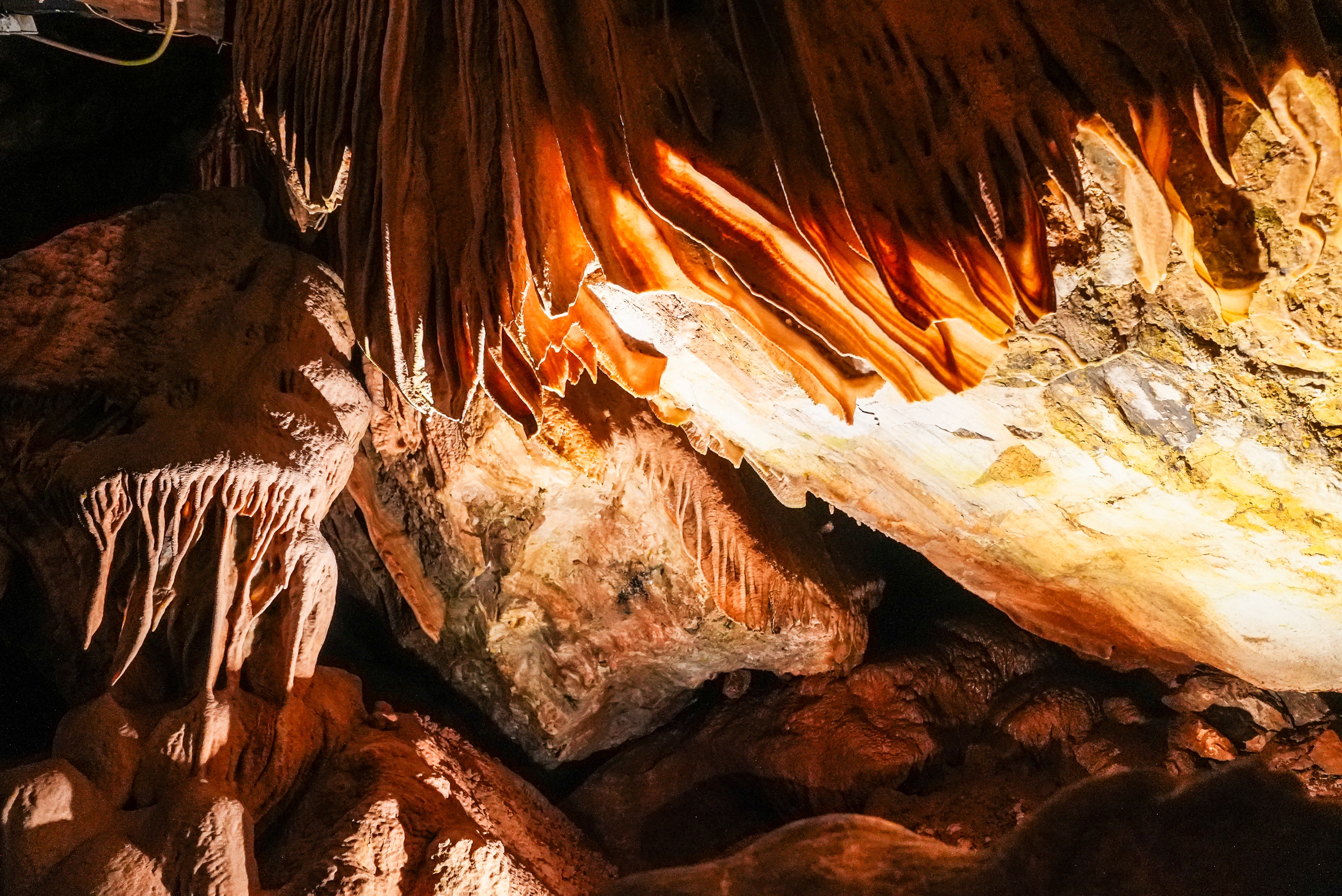 The image shows a cave interior with sharp, orange-brown stalactites hanging from the ceiling, illuminated by lights, creating a warm and textured ambiance.