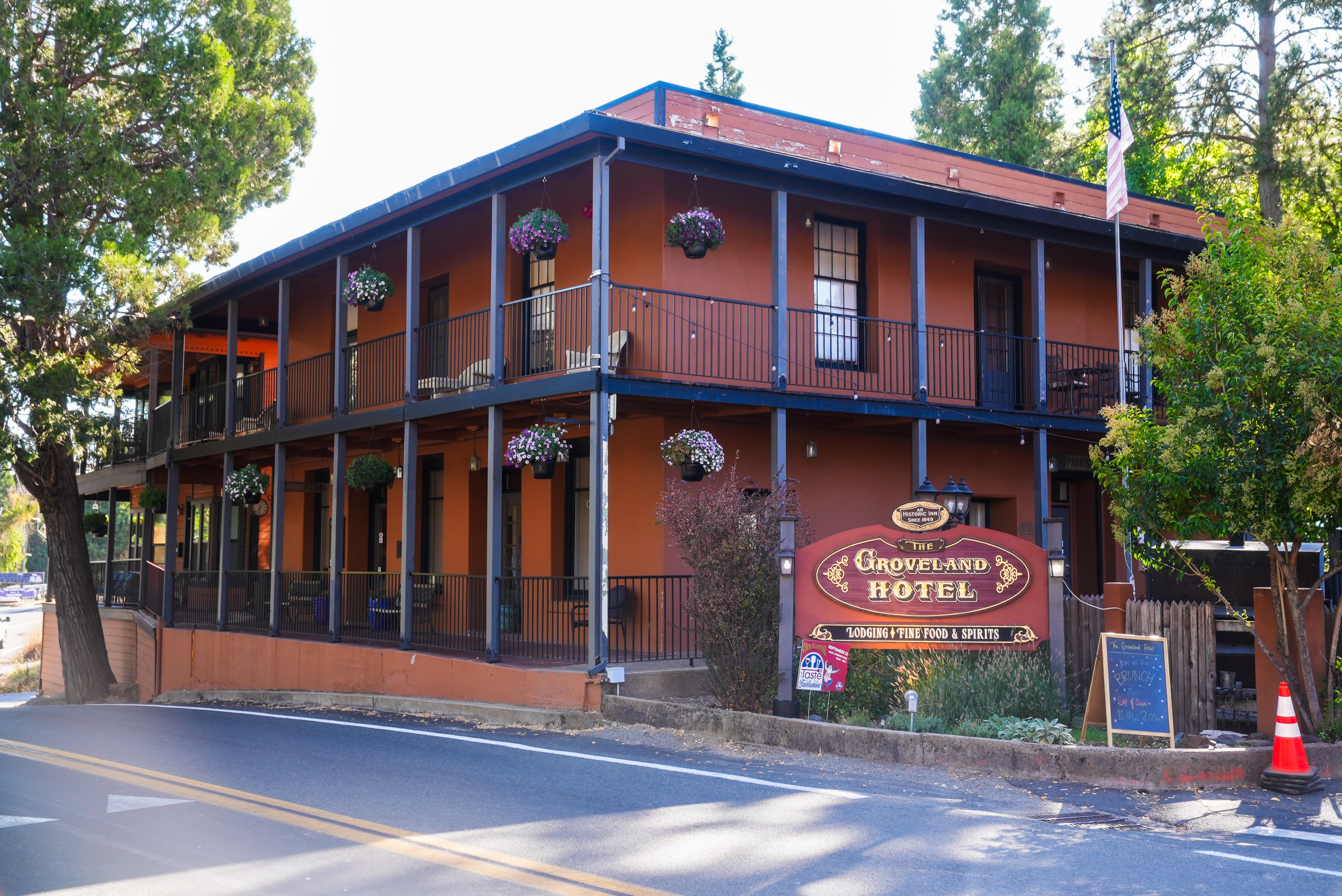 The image shows a two-story orange hotel with a covered balcony adorned with hanging plants. There is a sign reading &quot;Groveland Hotel&quot; by the roadside.