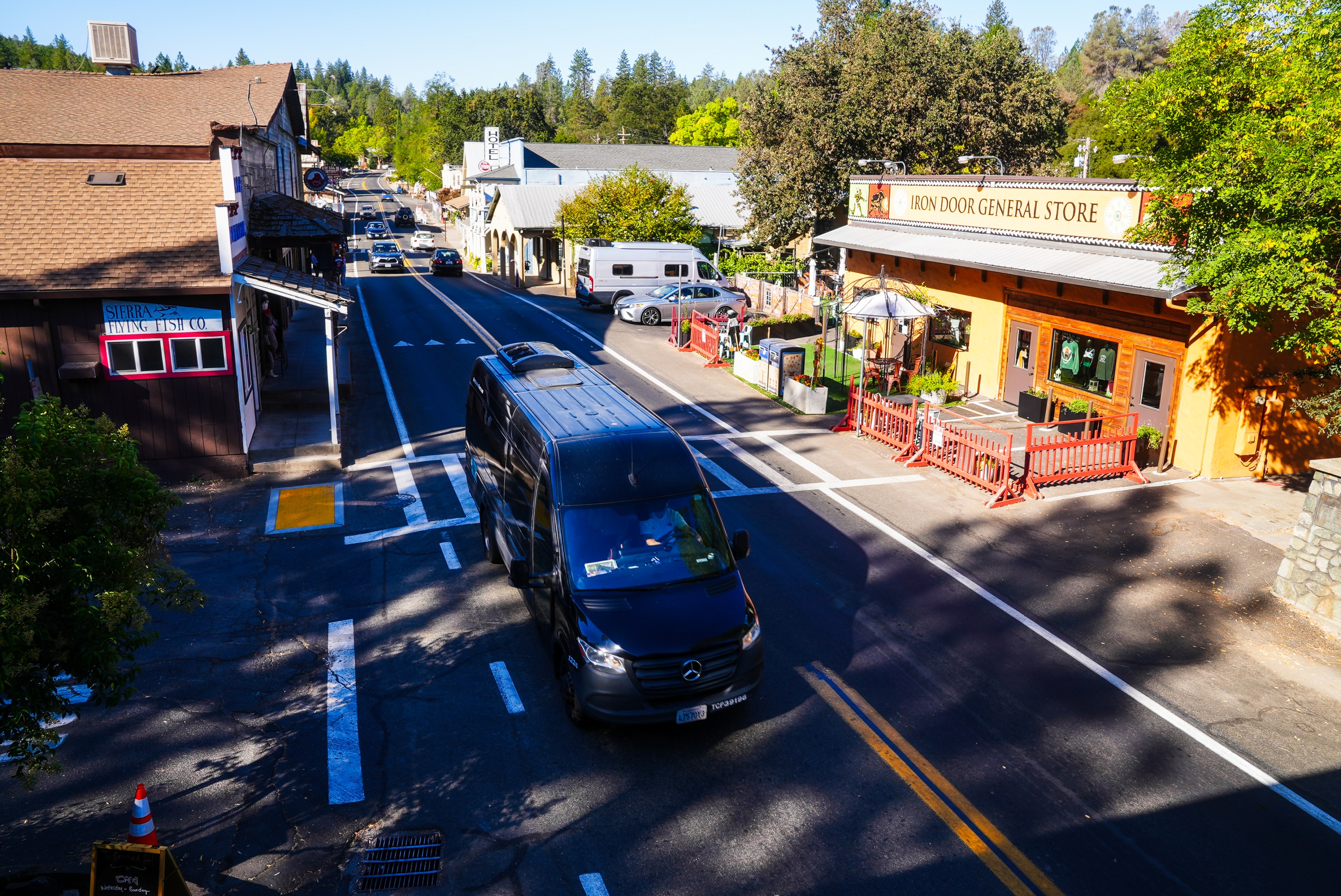 A small town street with parked cars, a black van, trees, and buildings, including a general store and a fishing company. The scene is sunny and quiet.