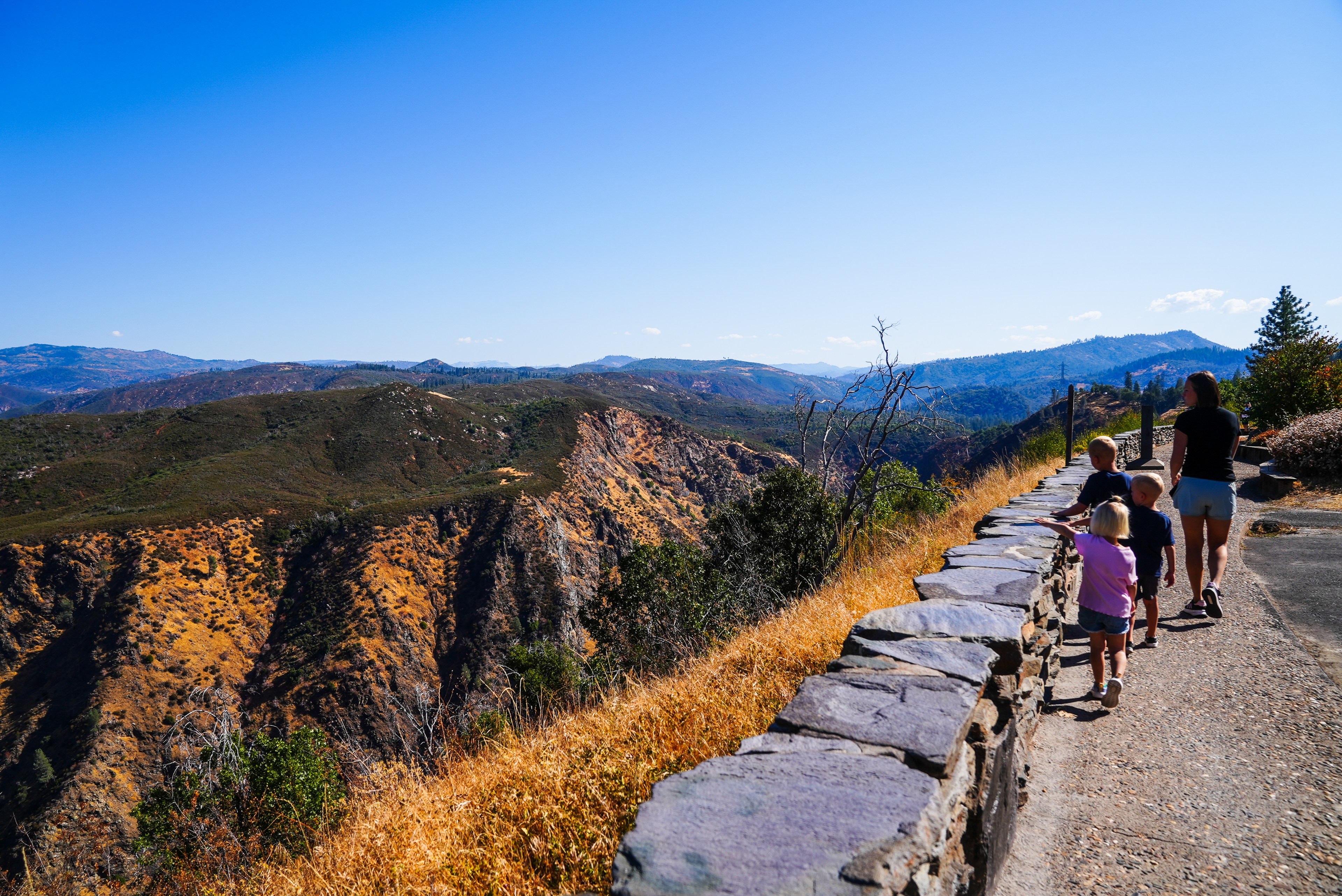 A woman and three children walk along a stone wall overlooking a rocky canyon, surrounded by dry grass and distant mountains under a clear blue sky.