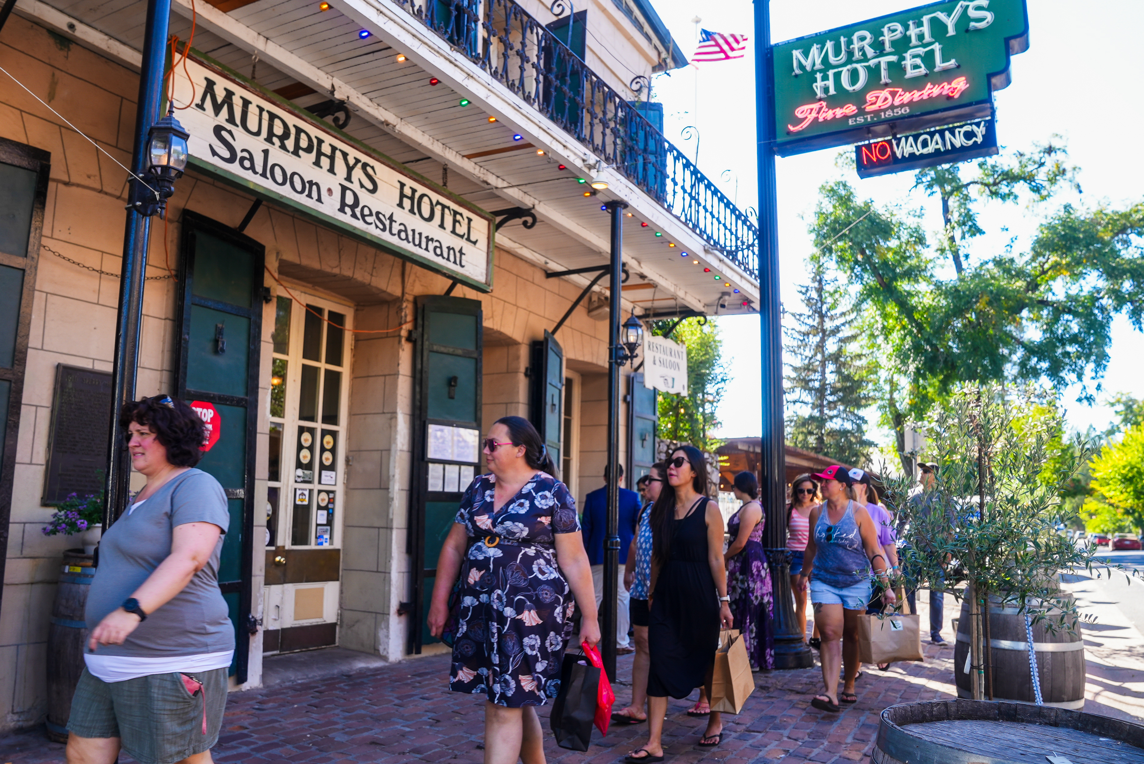 People walk along a lively street past Murphy's Hotel, with an American flag and colorful lights above. A &quot;No Vacancy&quot; sign is displayed, and trees line the background.