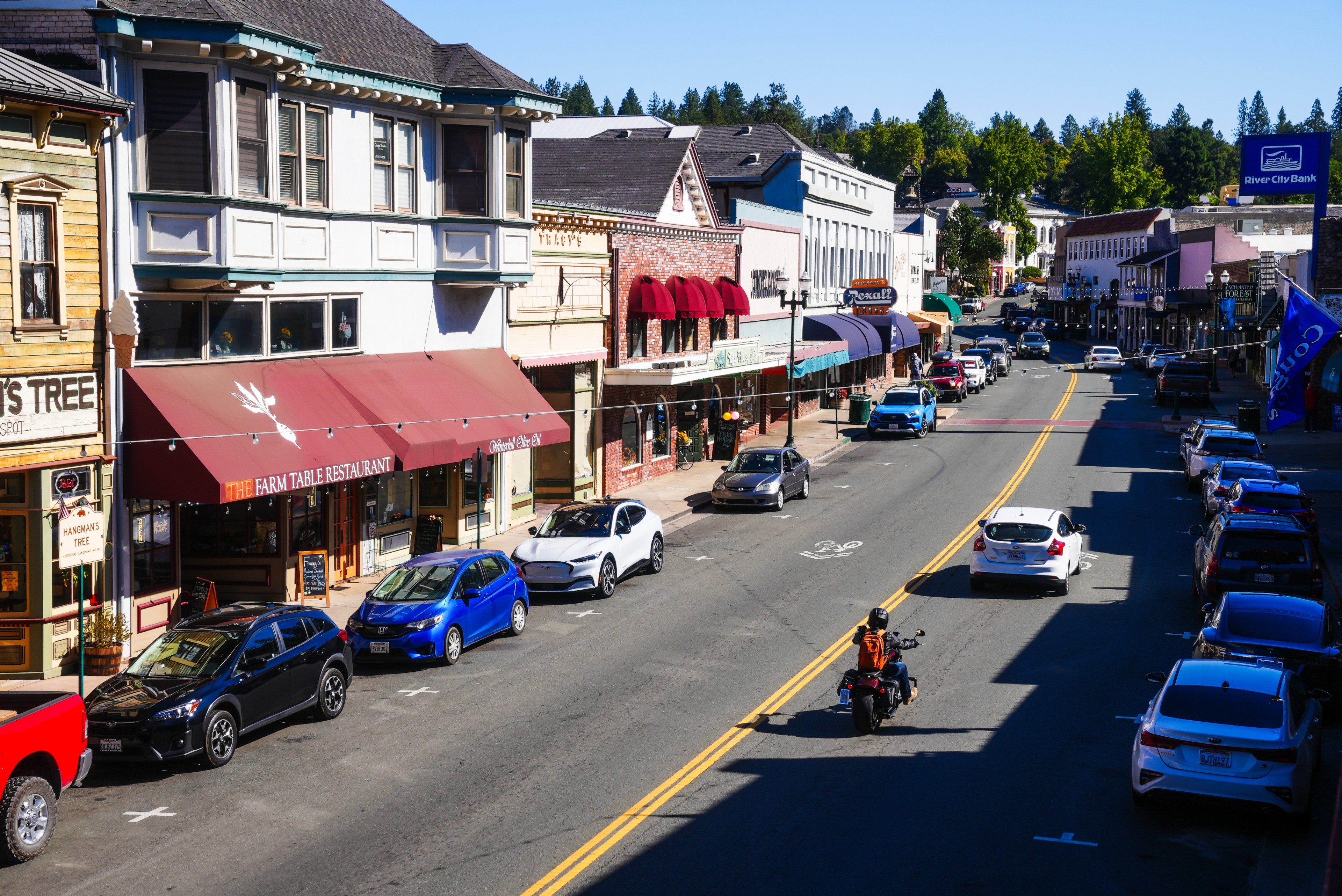 A bustling small-town street features shops with colorful awnings, parked cars, a motorcycle in motion, and a clear blue sky above.