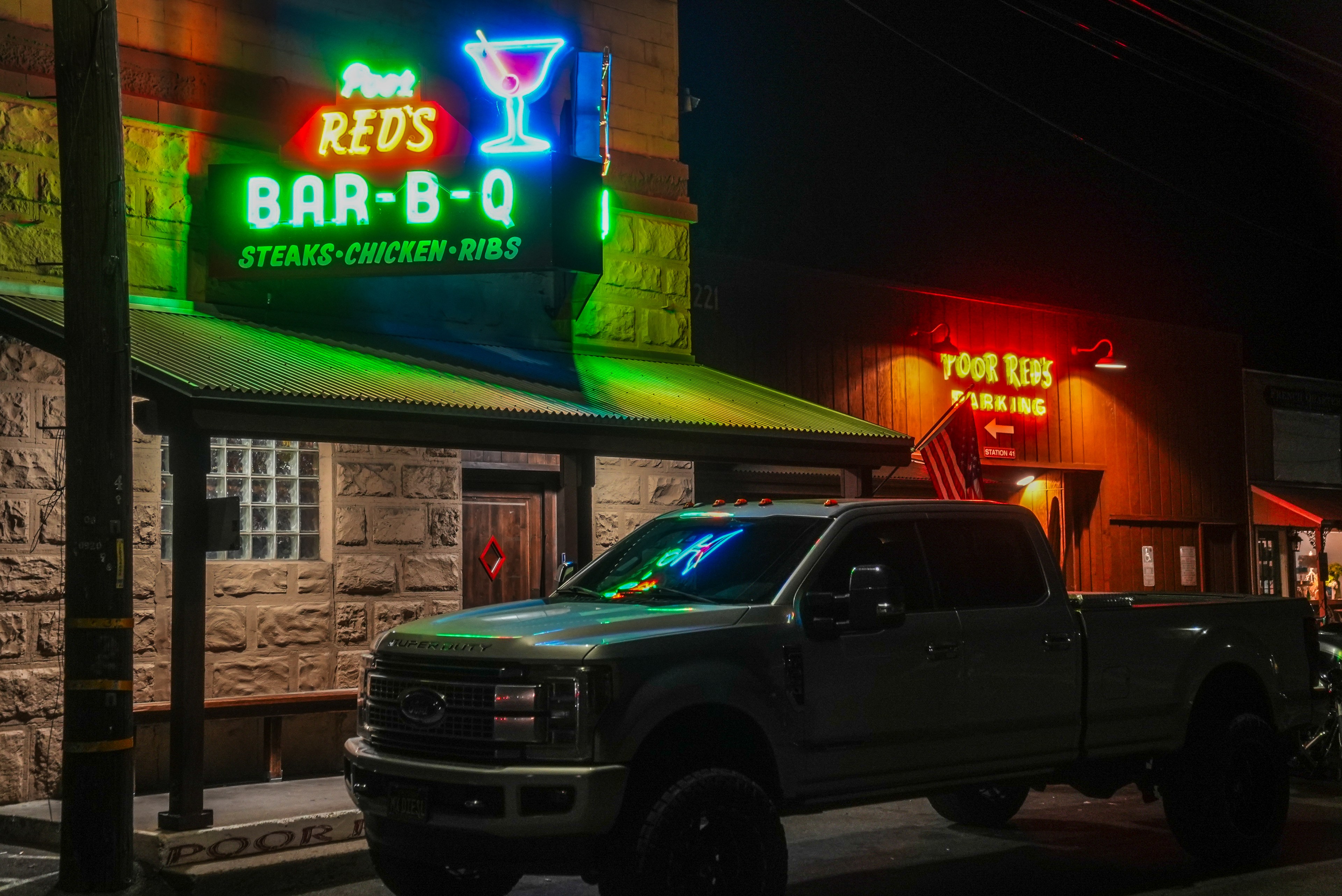 A neon-lit bar named Poor Red's shows its Bar-B-Q sign with a cocktail glass above a large truck parked in front at night.