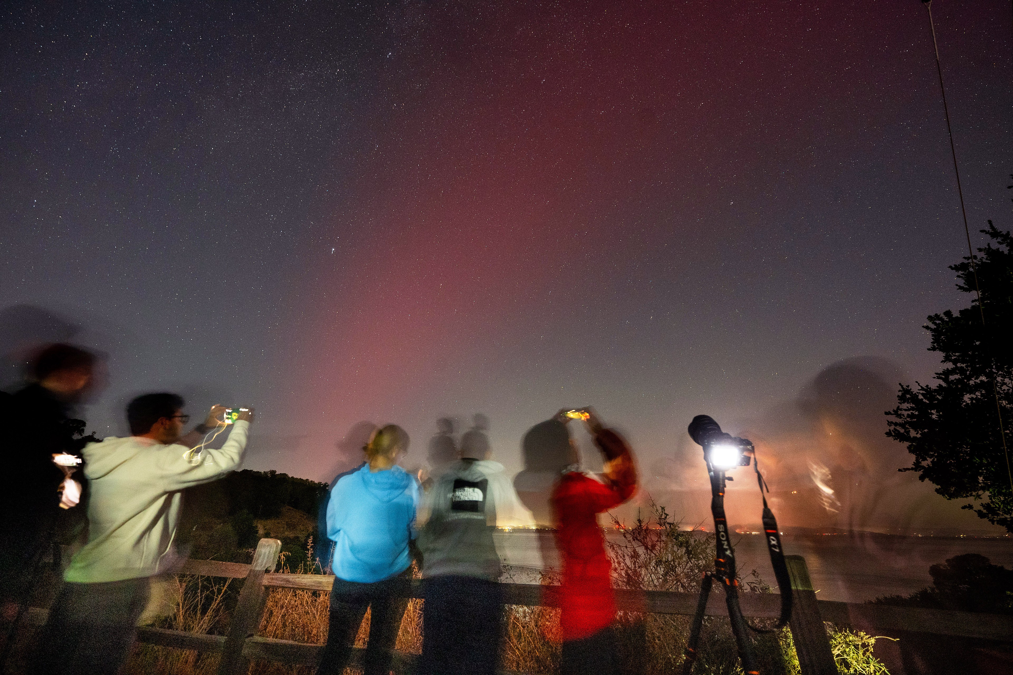 A group of blurry people stand by a wooden fence, photographing a night sky illuminated with faint red auroras. A tripod is set up, and trees are visible on the right.