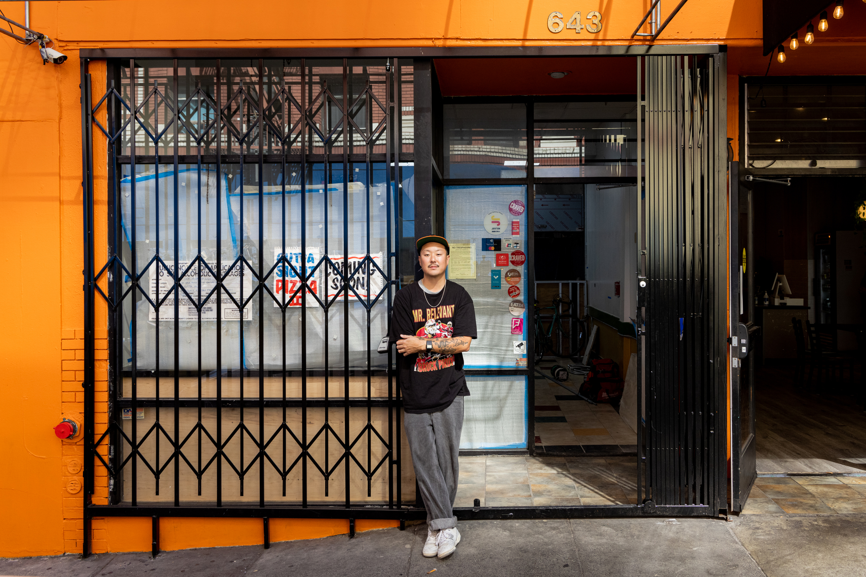 A person stands in front of an orange storefront with a metal gate. The door is open, and they are wearing casual clothes with arms crossed. The street number "643" is visible.