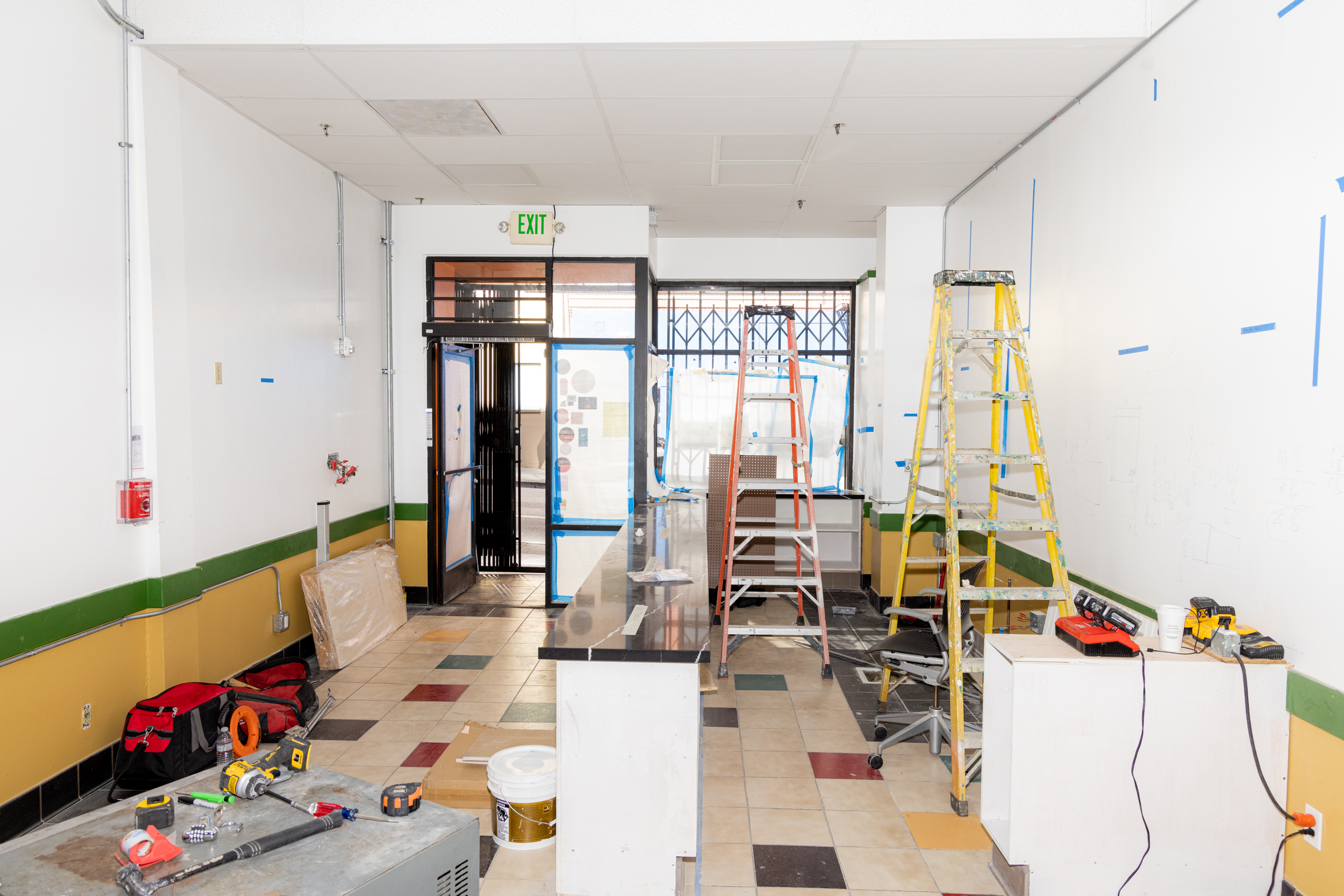 A room under renovation with ladders, tools, and construction materials scattered around. White walls and a tiled floor lead to a glass door marked &quot;EXIT.&quot;