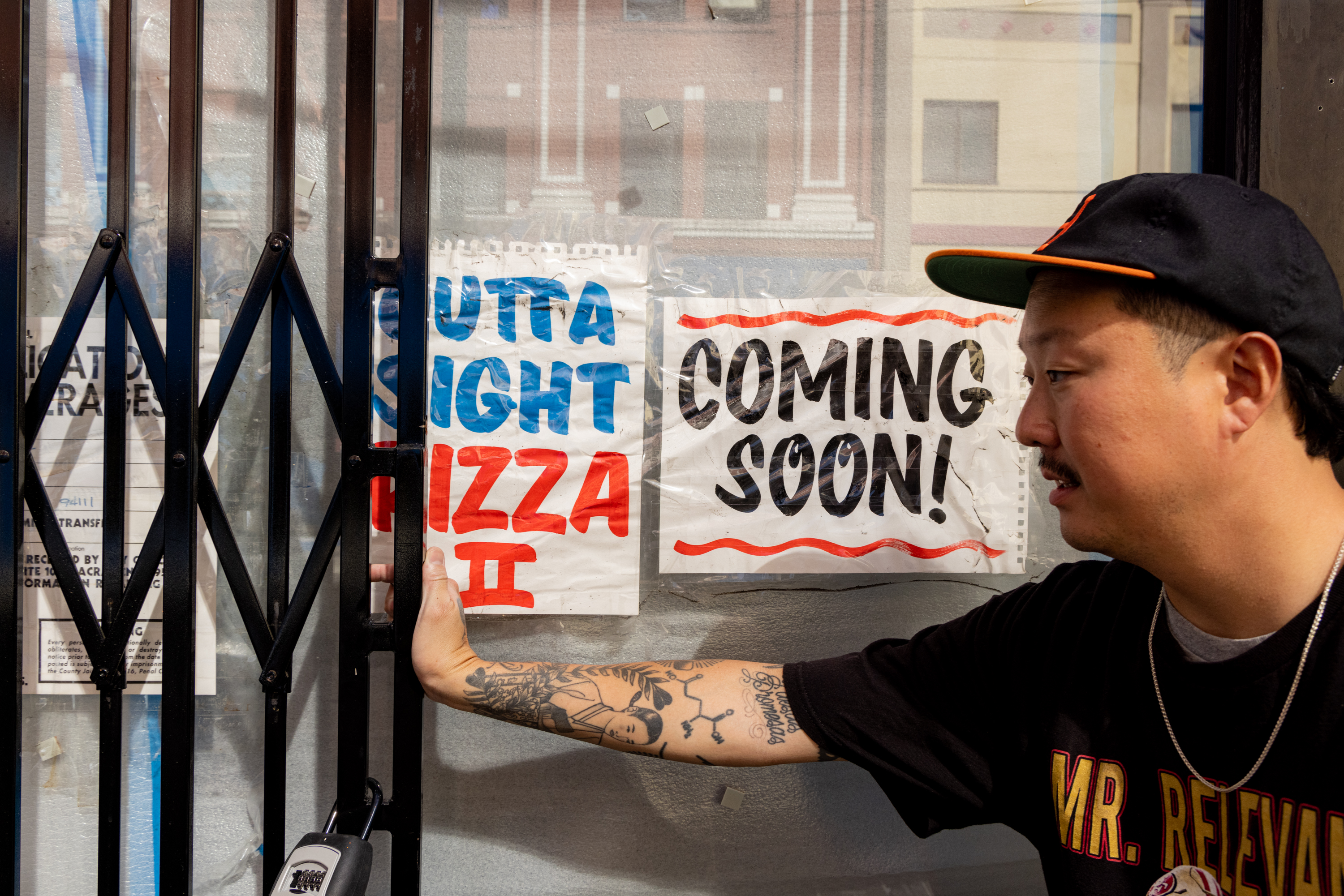 A man with tattoos holds a gate beside signs reading &quot;Outta Sight Pizza II&quot; and &quot;Coming Soon!&quot; on a window. He's wearing a black cap and a &quot;Mr. Relevant&quot; shirt.