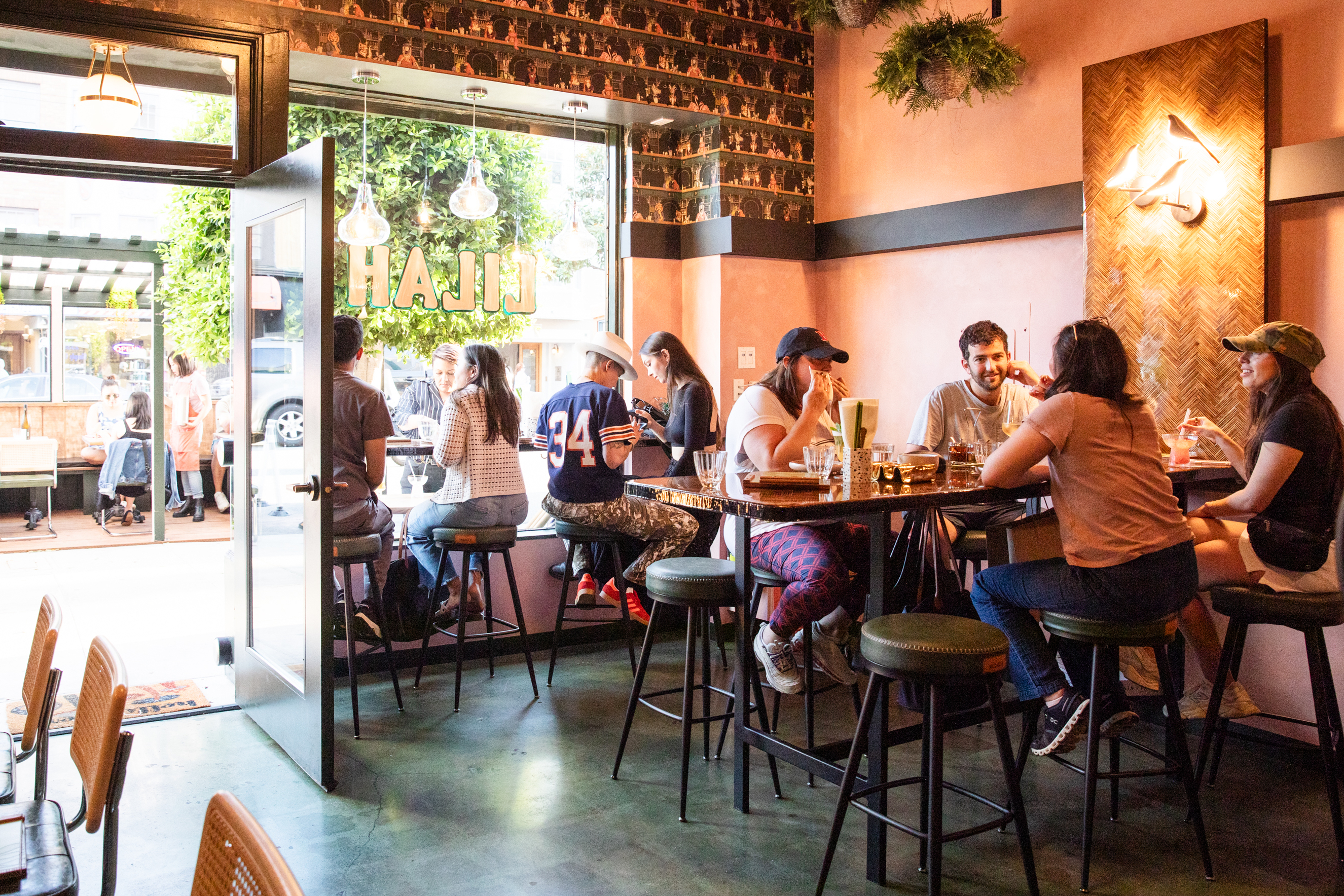 People are sitting at high tables inside a cozy café with a warm ambiance. A few are near the open door enjoying drinks, and there’s greenery outside.