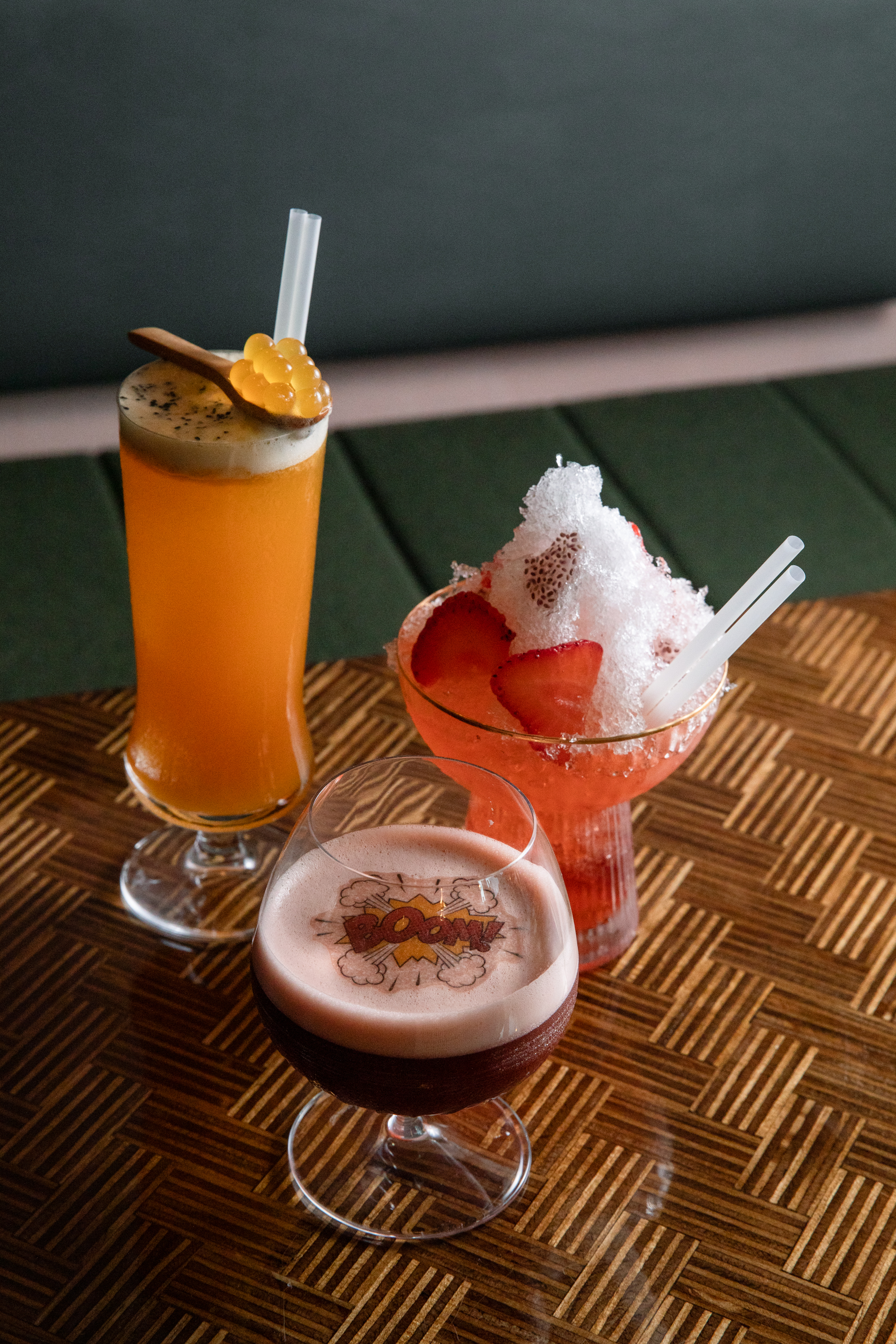 Three colorful drinks sit on a wooden table: an orange cocktail with a garnish, a red shaved ice drink with strawberries, and a foamy dark drink with &quot;BOOM!&quot; on top.