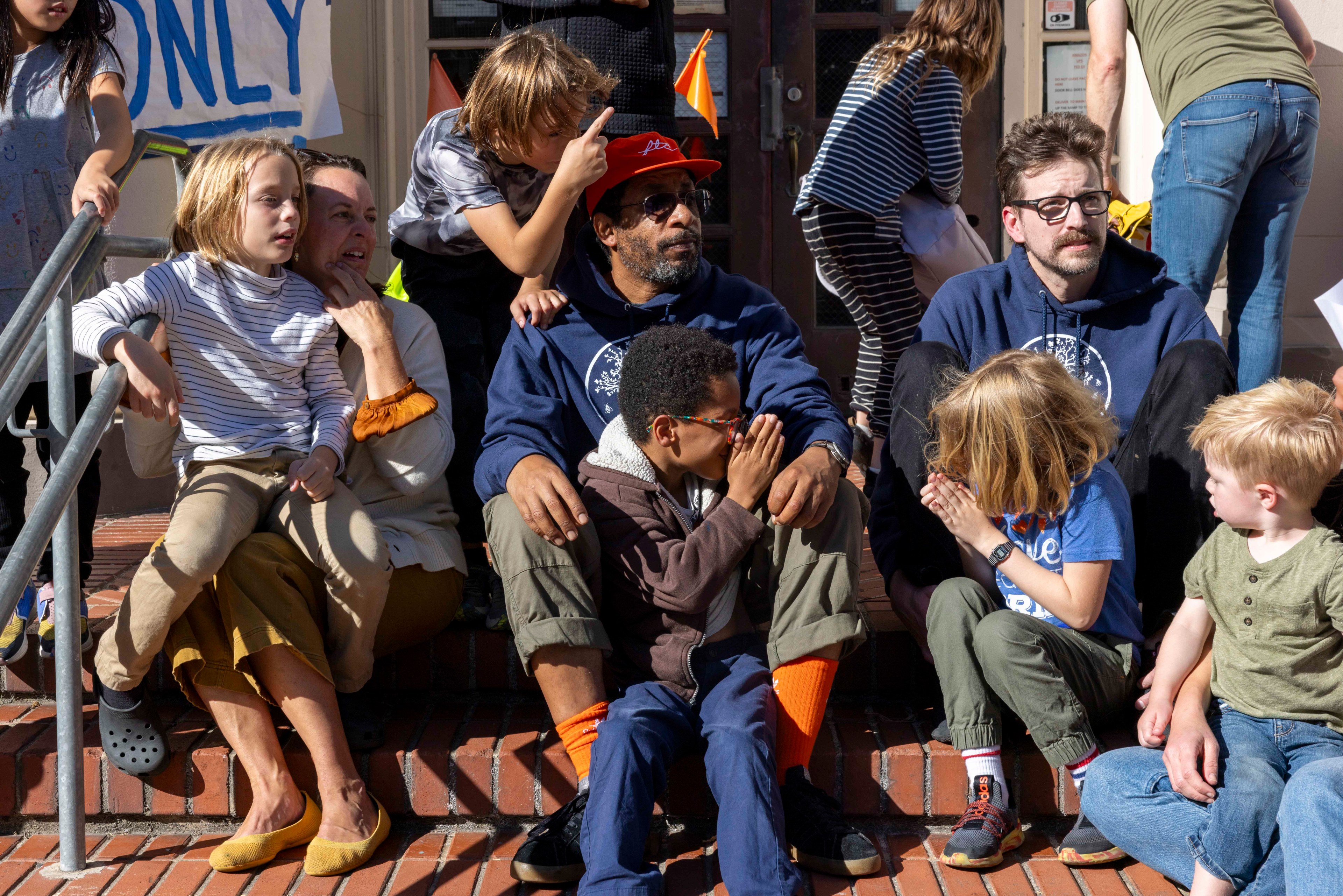 A group of people, including children and adults, sit on outdoor steps, talking and smiling. A person in the center wears a red cap and sunglasses.