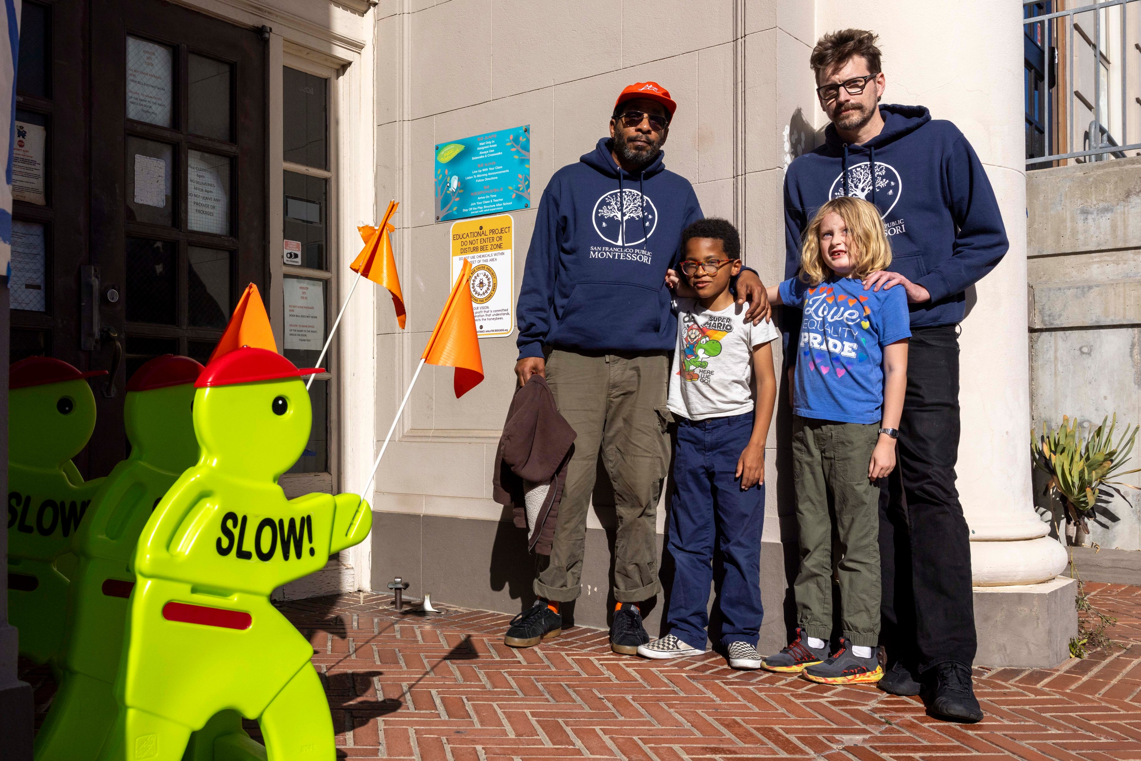 Two adults and two kids stand outside a building with &quot;SLOW!&quot; signs nearby. The adults wear blue hoodies; the kids wear graphic tees. It's a sunny day.