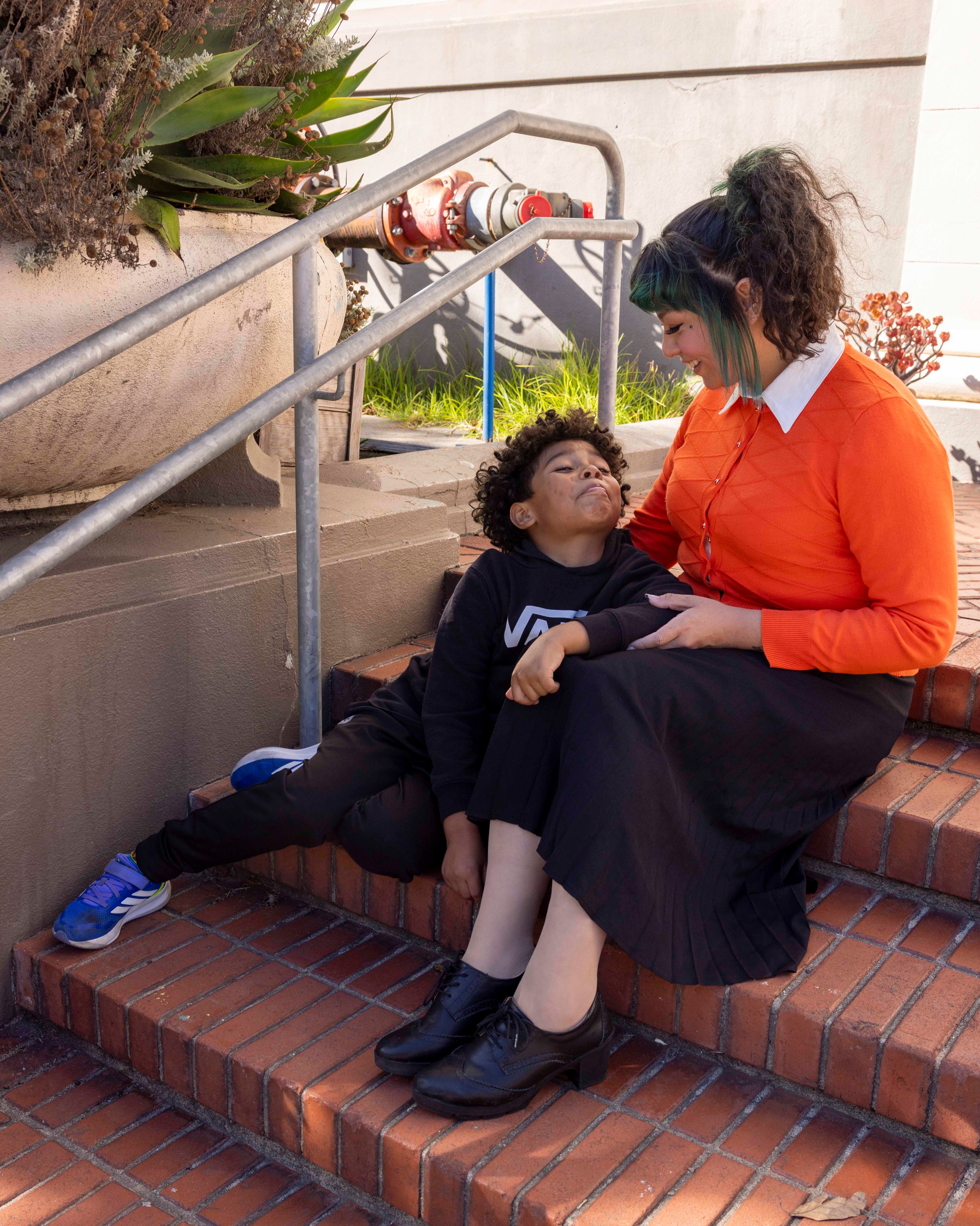 A child and an adult sit on brick steps, smiling at each other. The adult wears an orange sweater and black skirt, while the child sports a black outfit and blue shoes.