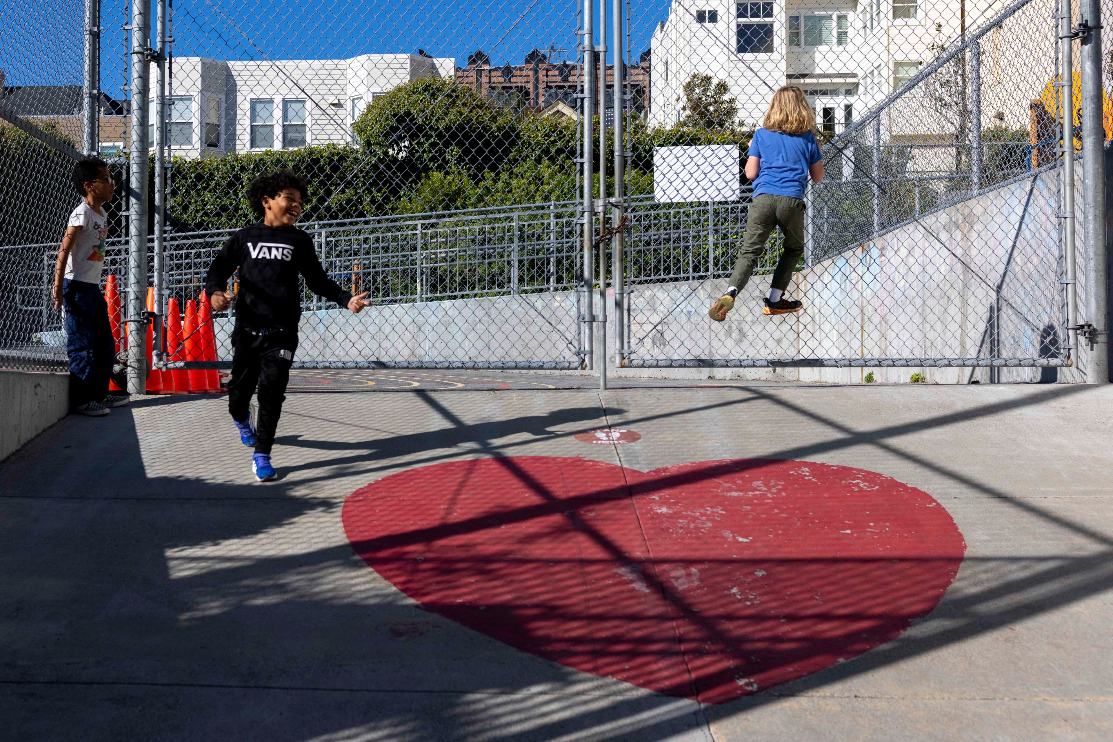 Children play near a chain-link fence, with one climbing it. A large red heart is painted on the ground, and city buildings are visible in the background.