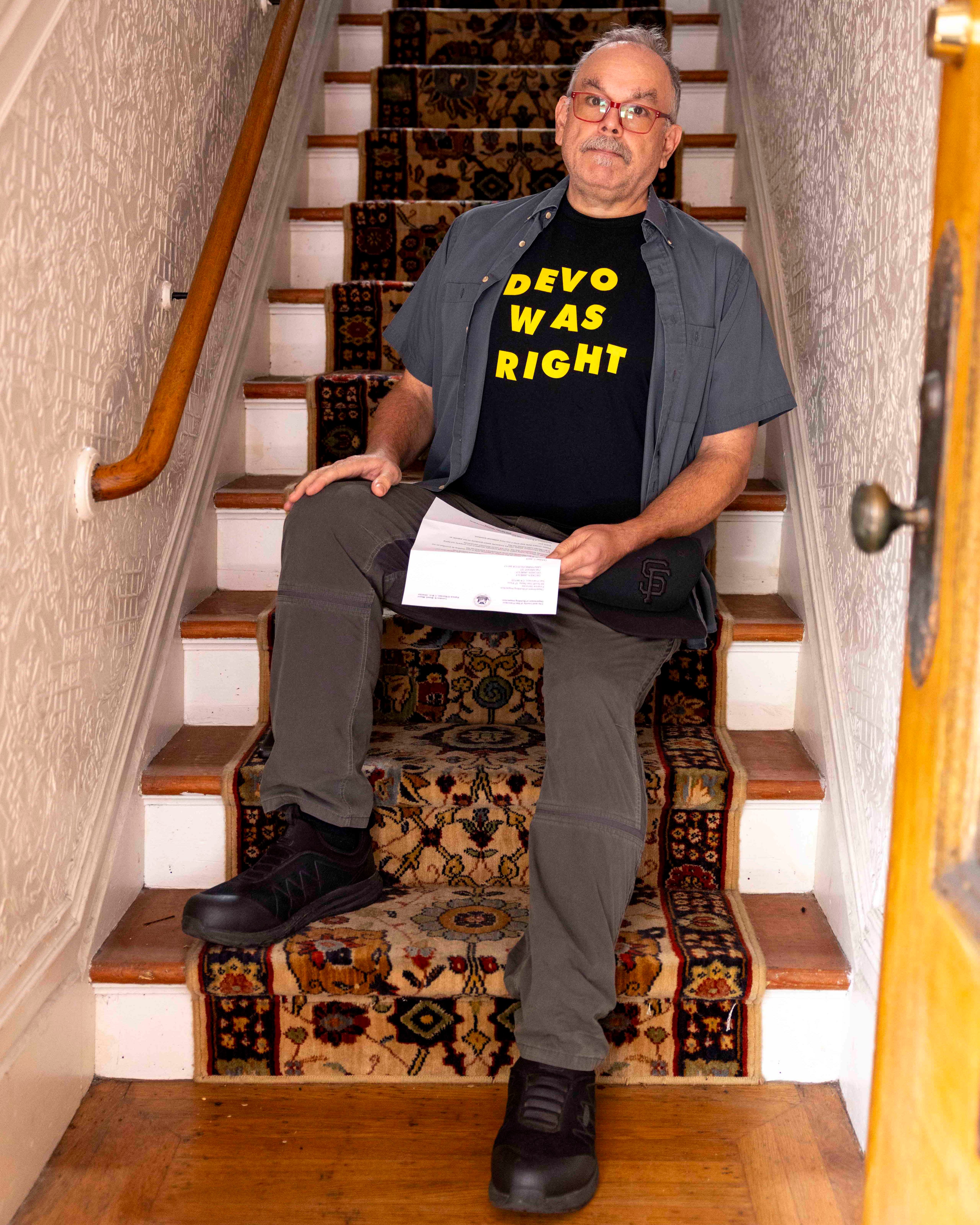 A person sits on a patterned staircase wearing a "DEVO WAS RIGHT" t-shirt, gray pants, and black shoes, holding a piece of paper, with a wooden handrail beside them.