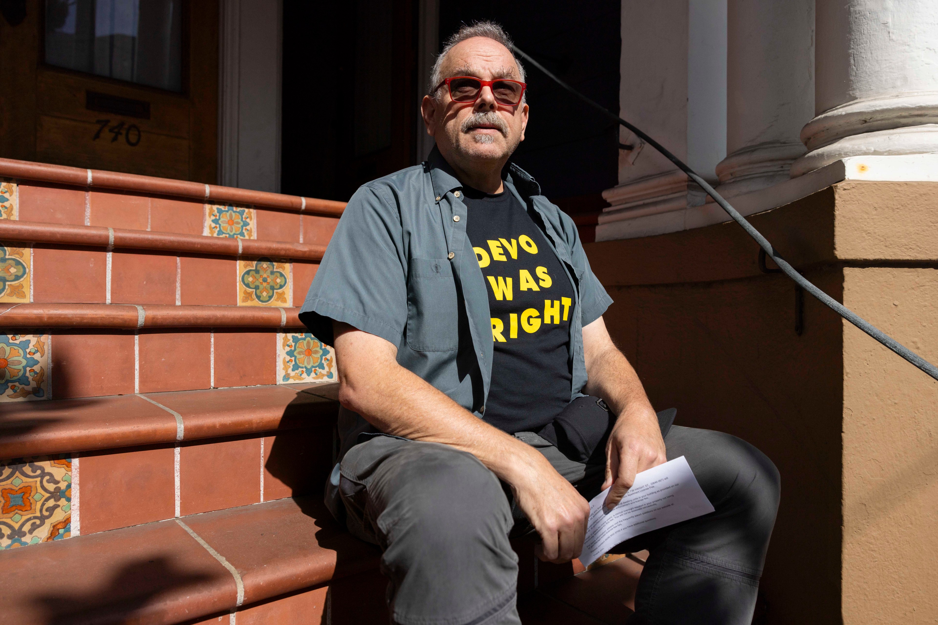 A man in sunglasses sits on decorative tiled steps, wearing a shirt saying &quot;DEVO WAS RIGHT.&quot; He holds a paper and is wearing casual clothing.