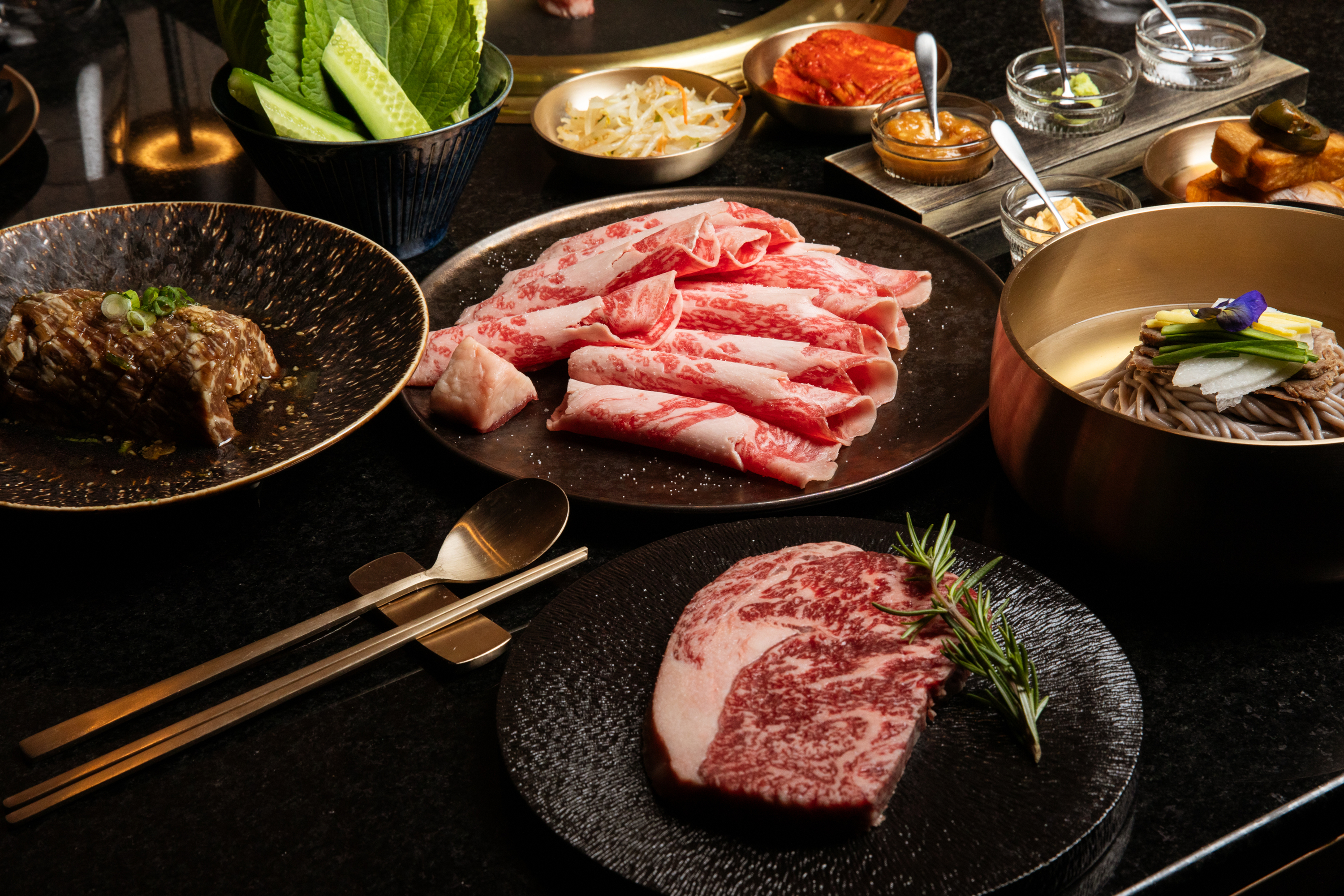 Slices of marbled beef and a steak are arranged on plates. There are soba noodles, leafy greens, and small bowls of sauces and pickles on a dark table.