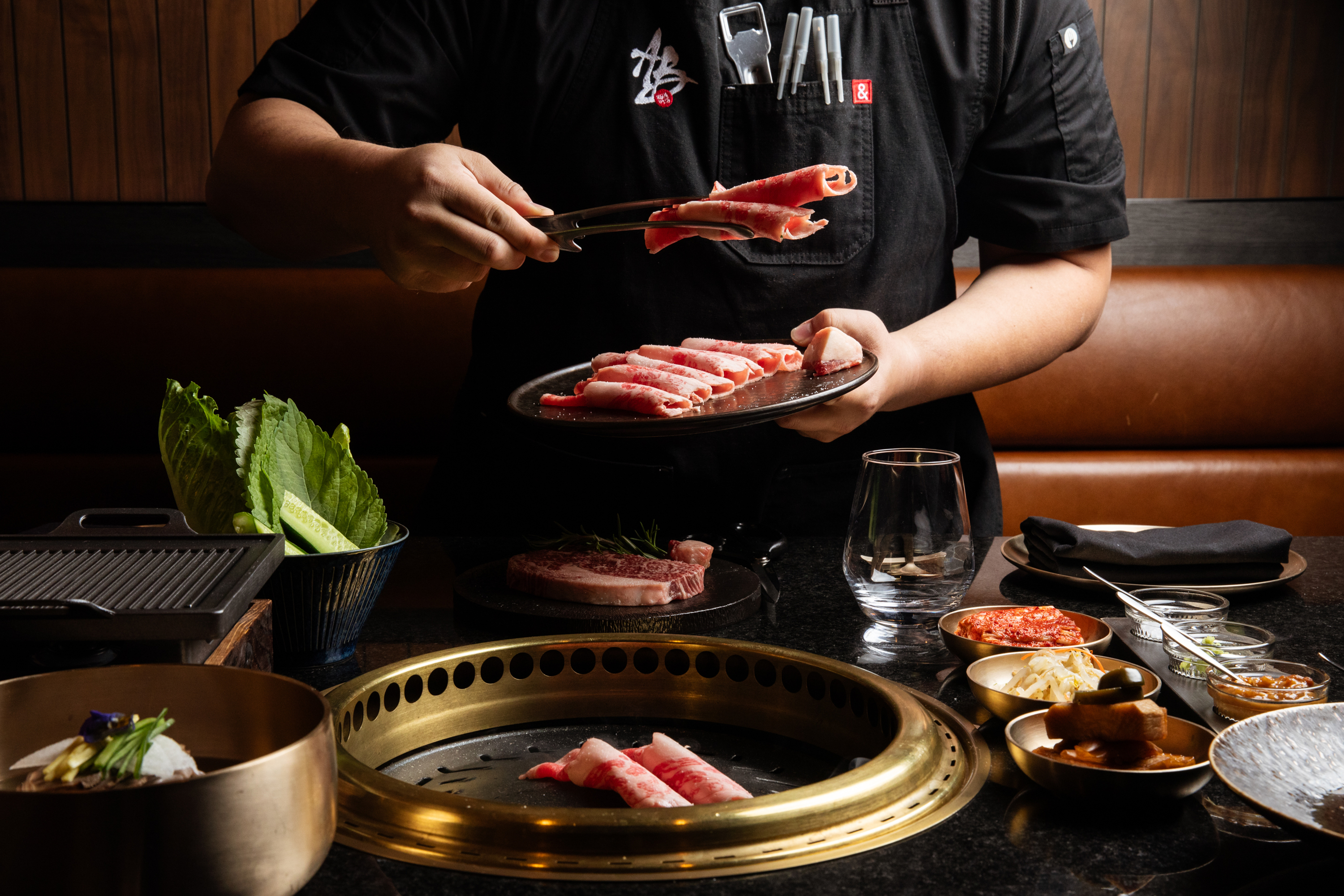 A person is grilling meat at a table with a round grill. Various dishes, including lettuce and side items, are displayed around them, creating a cozy dining setting.