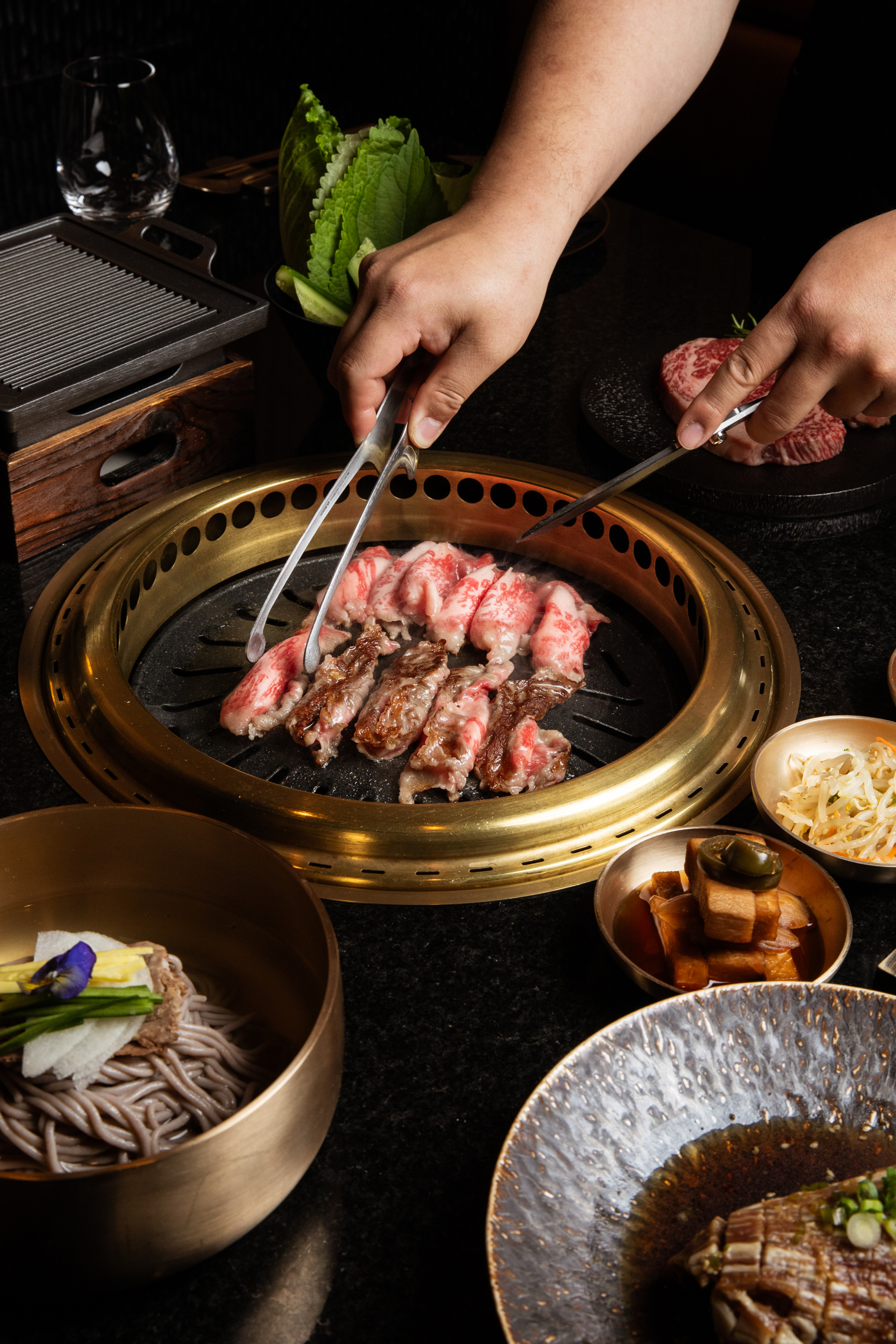 Hands grill marbled beef on a circular grill, surrounded by dishes of noodles, greens, pickled vegetables, and sauces.