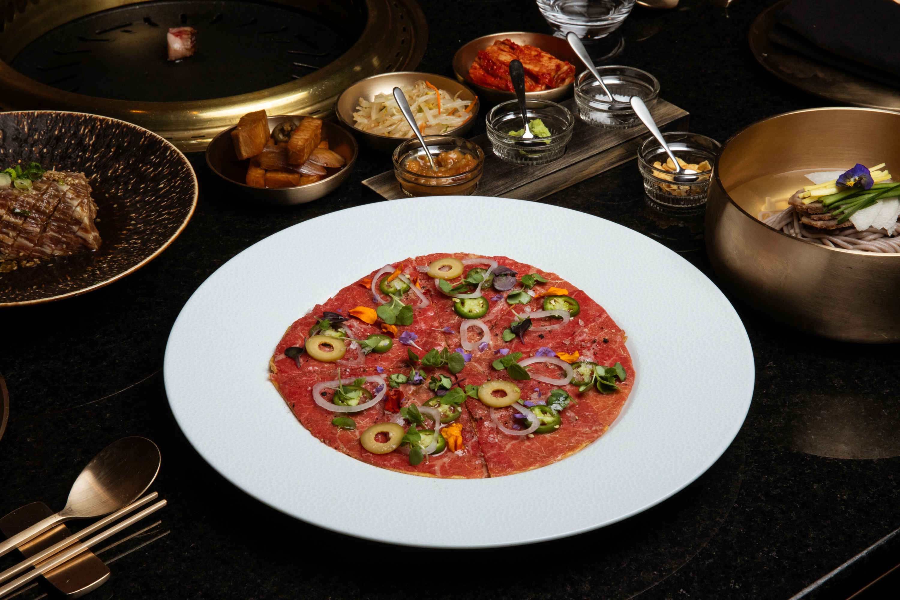 A plate of thinly sliced beef topped with olives, onion, and herbs is surrounded by various side dishes like kimchi, soba noodles, and sauces on a dark table.