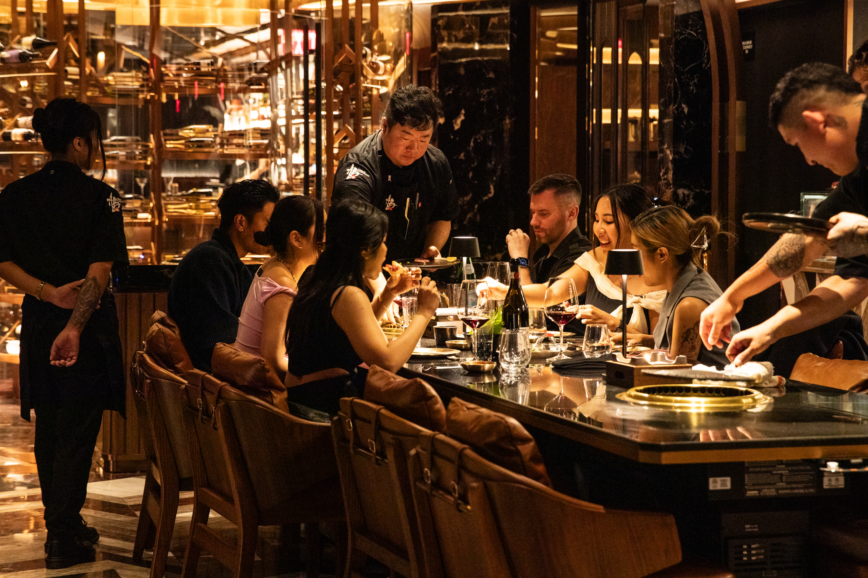 A group of people is dining at an elegant restaurant with a modern, warm ambiance. Chefs are serving food, and the table is set with wine glasses and various dishes.