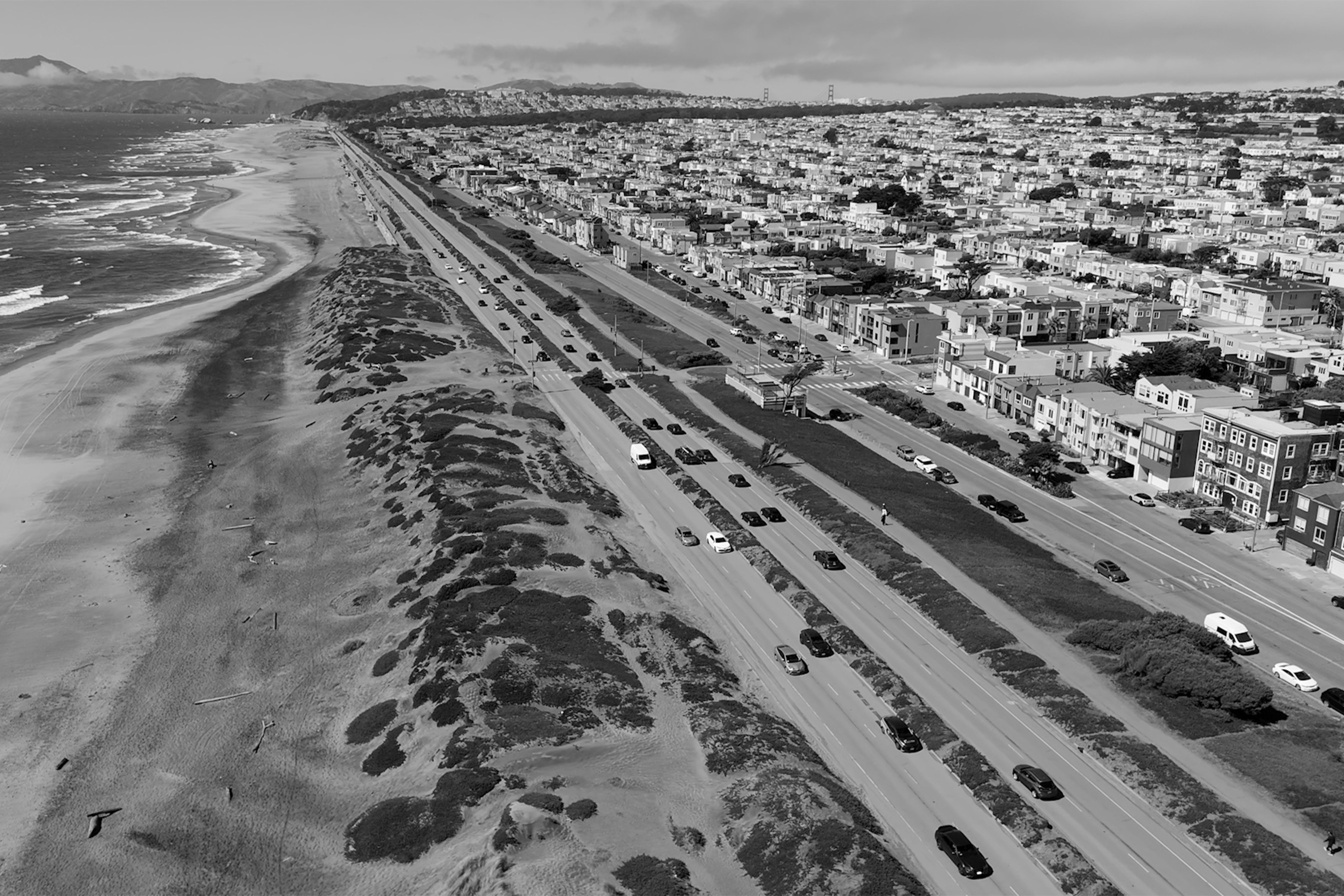 A coastal scene shows a long beach with waves on the left and a busy road bordered by houses on the right. The area is bustling with traffic.