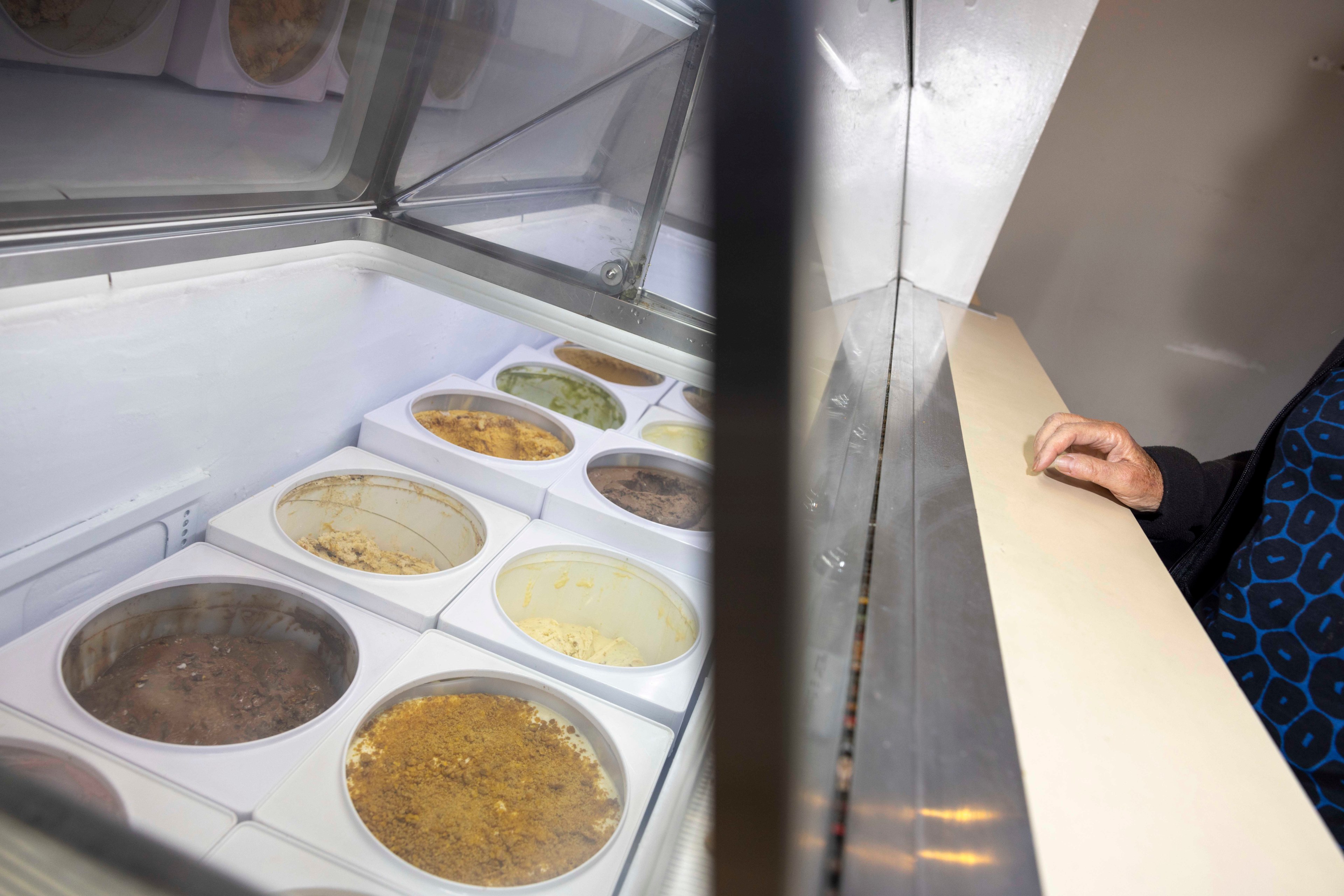 An ice cream shop display shows various flavors in tubs. A customer's hand rests on the glass counter, partially visible on the right side.