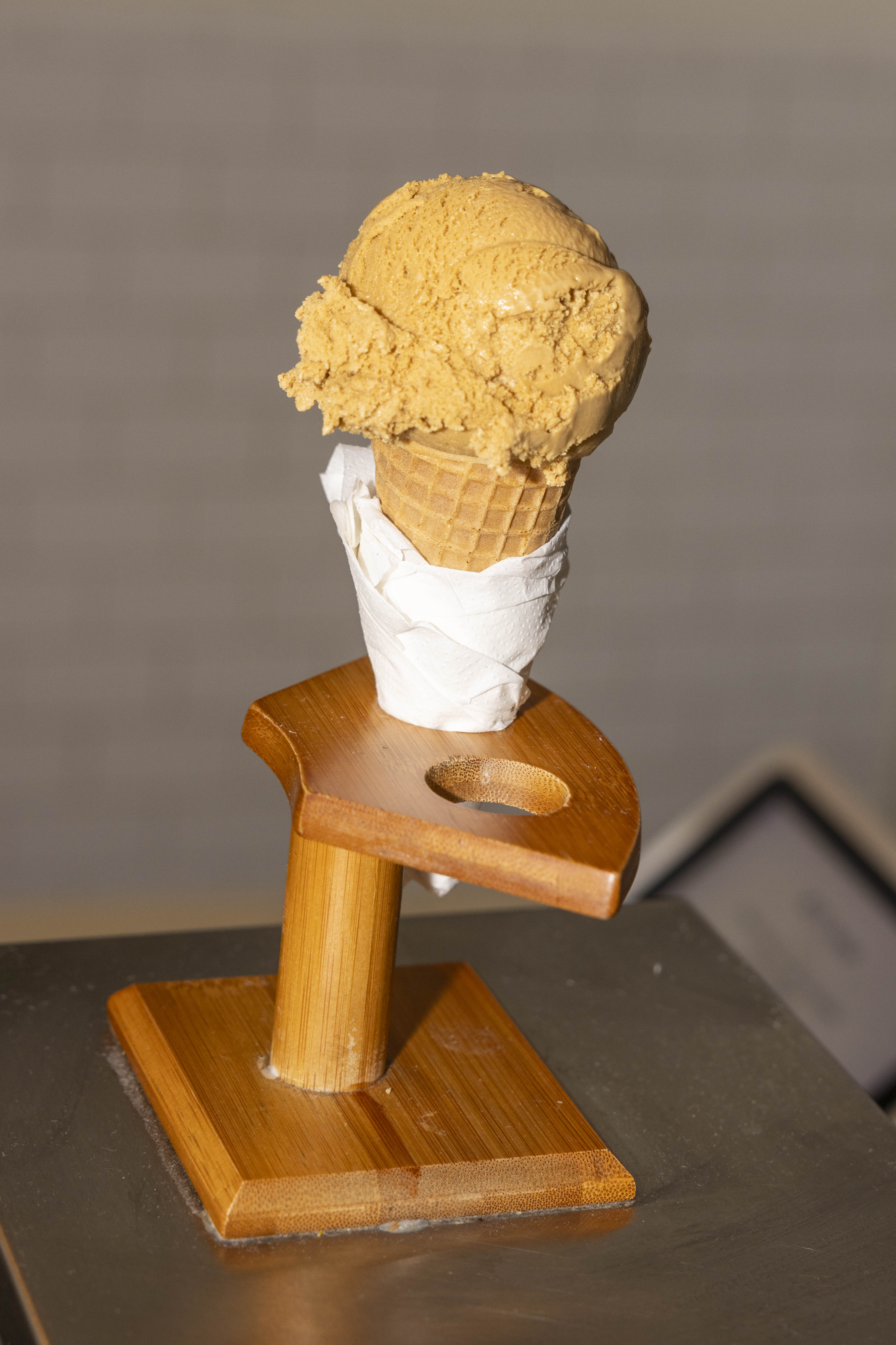 An ice cream cone with a scoop of caramel-colored ice cream is displayed on a wooden stand, wrapped neatly with a napkin.