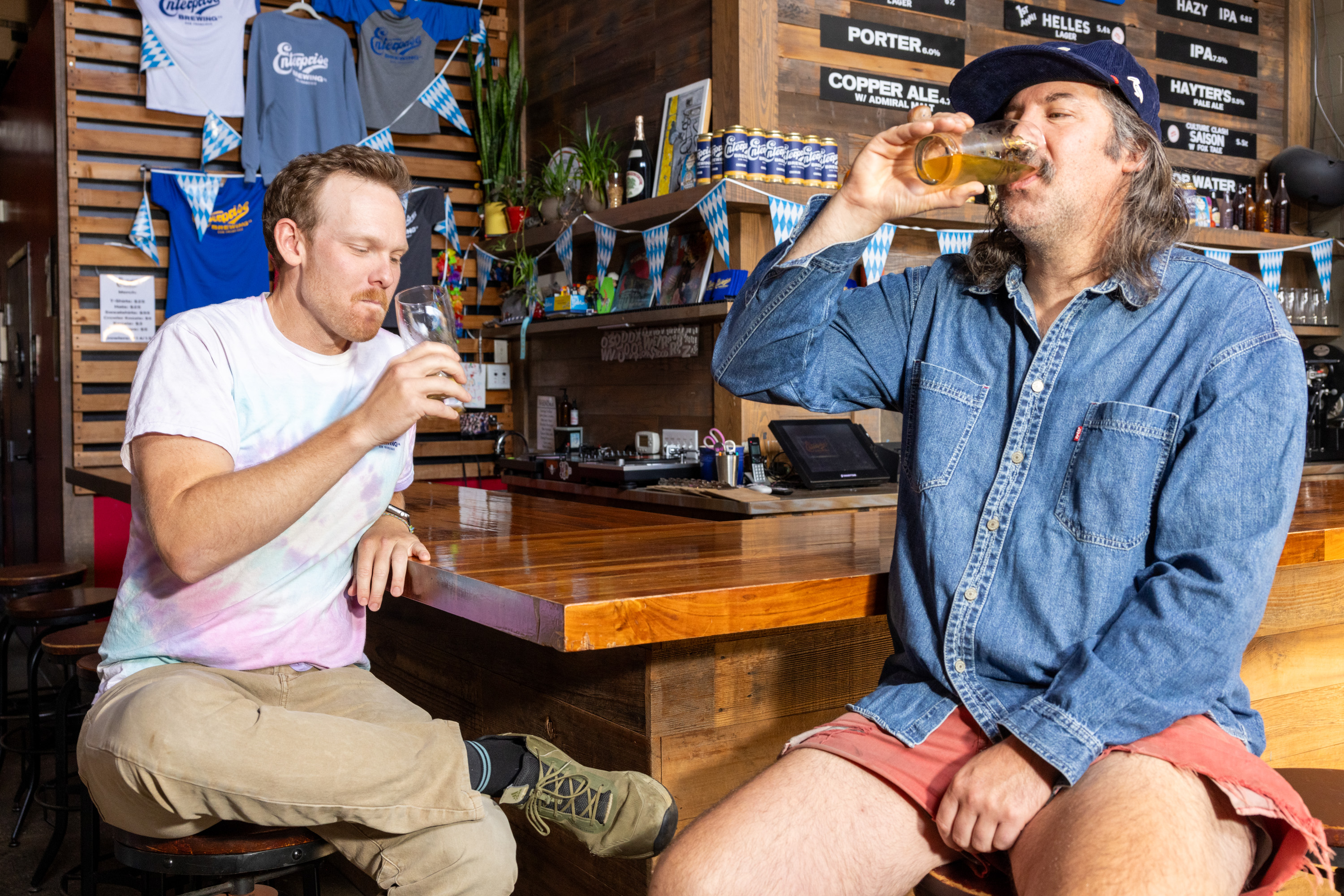 Two men sit at a wooden bar, each drinking beer. One wears a tie-dye shirt, the other a denim shirt with a cap. Beer-related decor hangs around them.