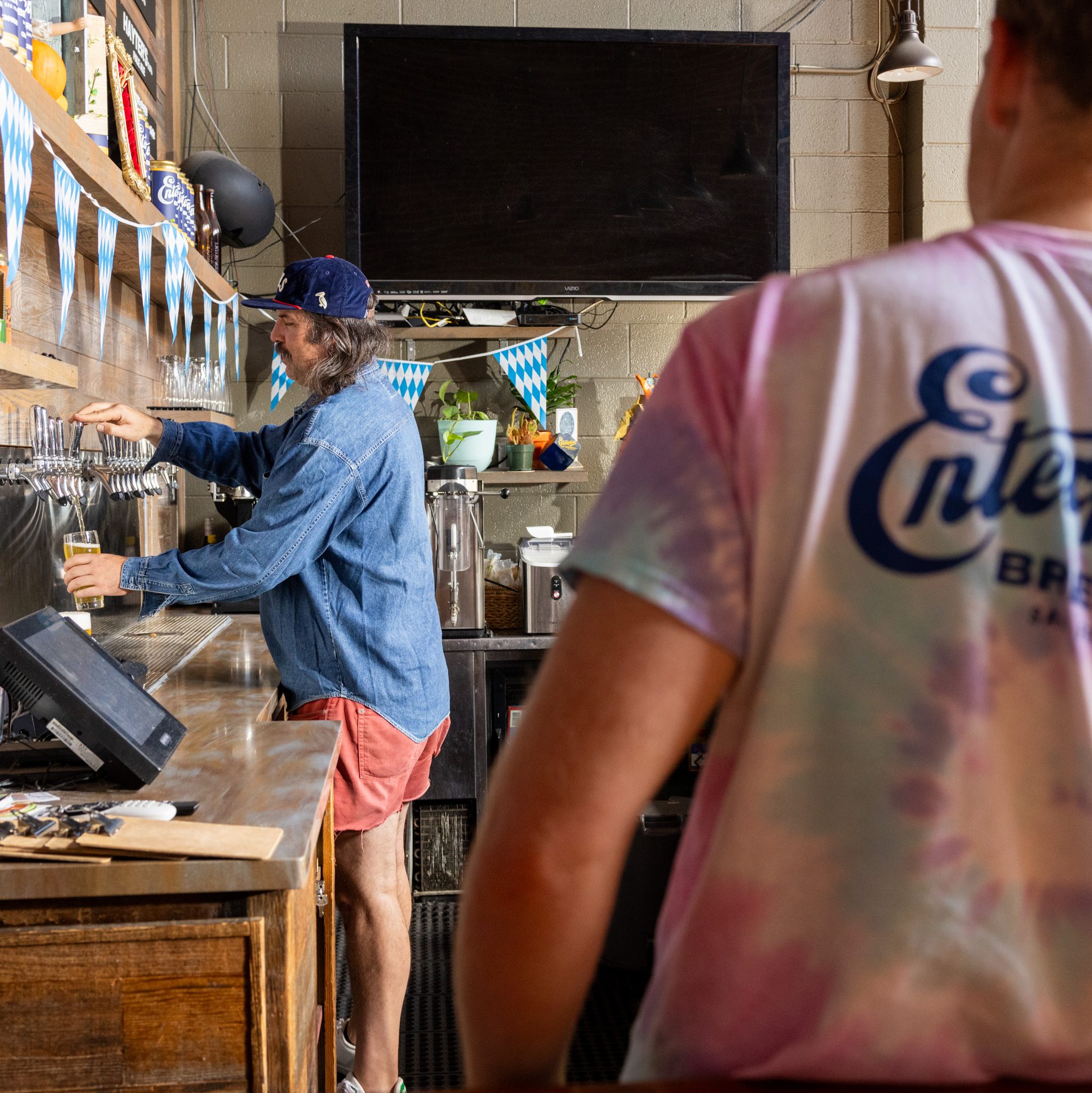 A man in a cap and denim shirt pours a drink from taps behind a bar, decorated with blue and white bunting. Another person in a tie-dye shirt stands nearby.