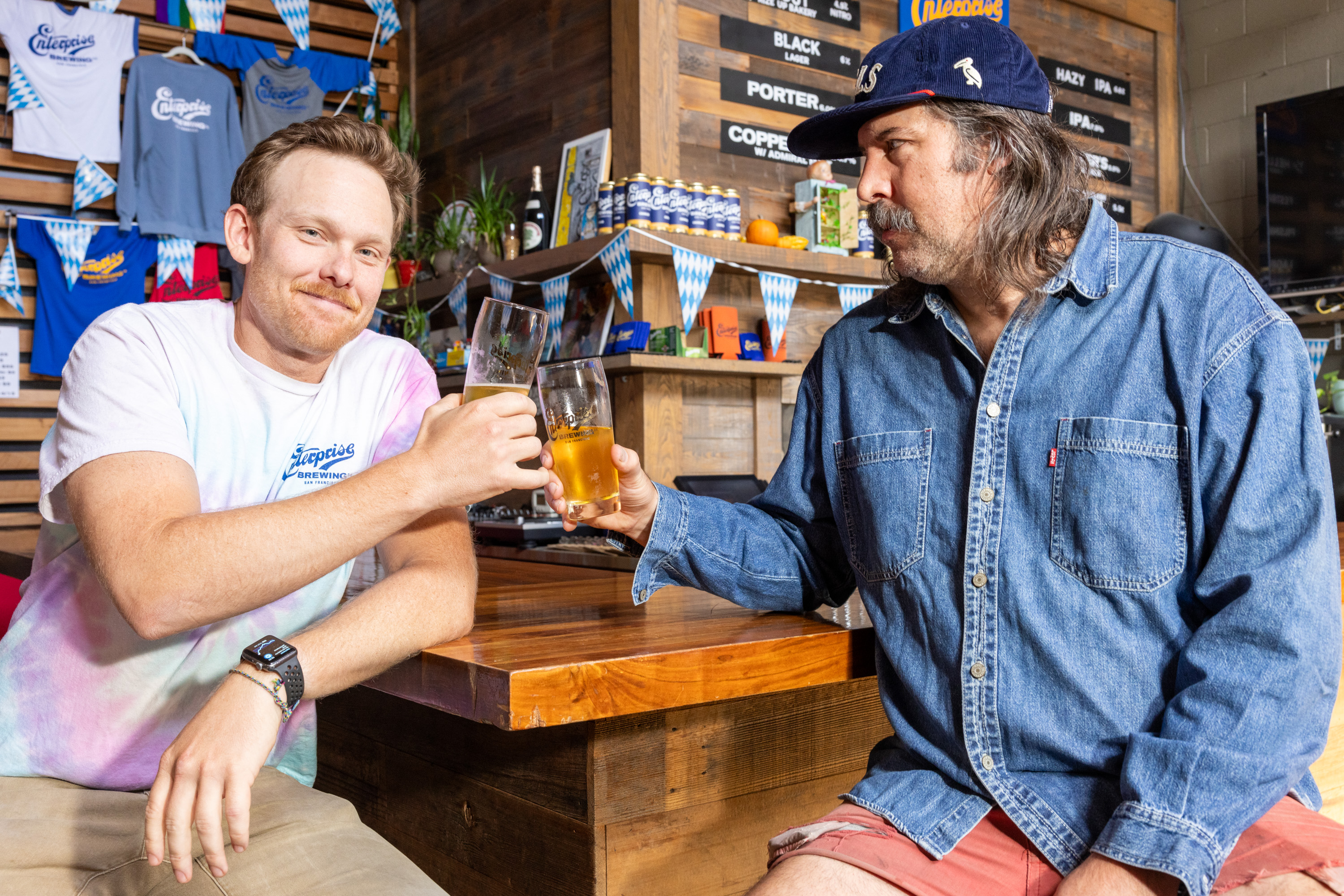 Two men are sitting at a wooden bar, smiling, and clinking their glasses of beer. The background features brewery merchandise and decor.