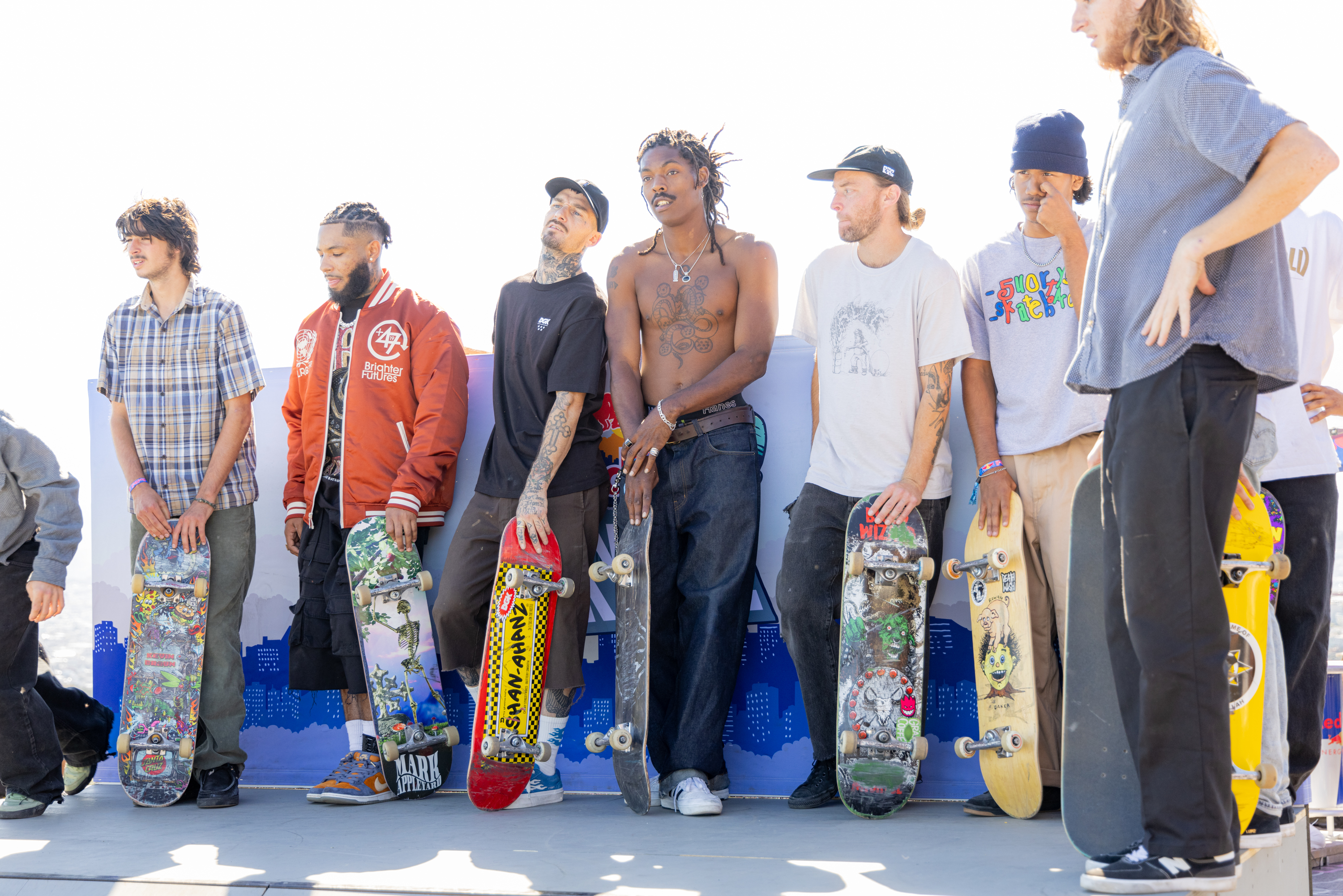 A group of men stand in a row, each holding a skateboard. They appear relaxed and casual, with tattoos visible on some, and different clothing styles.