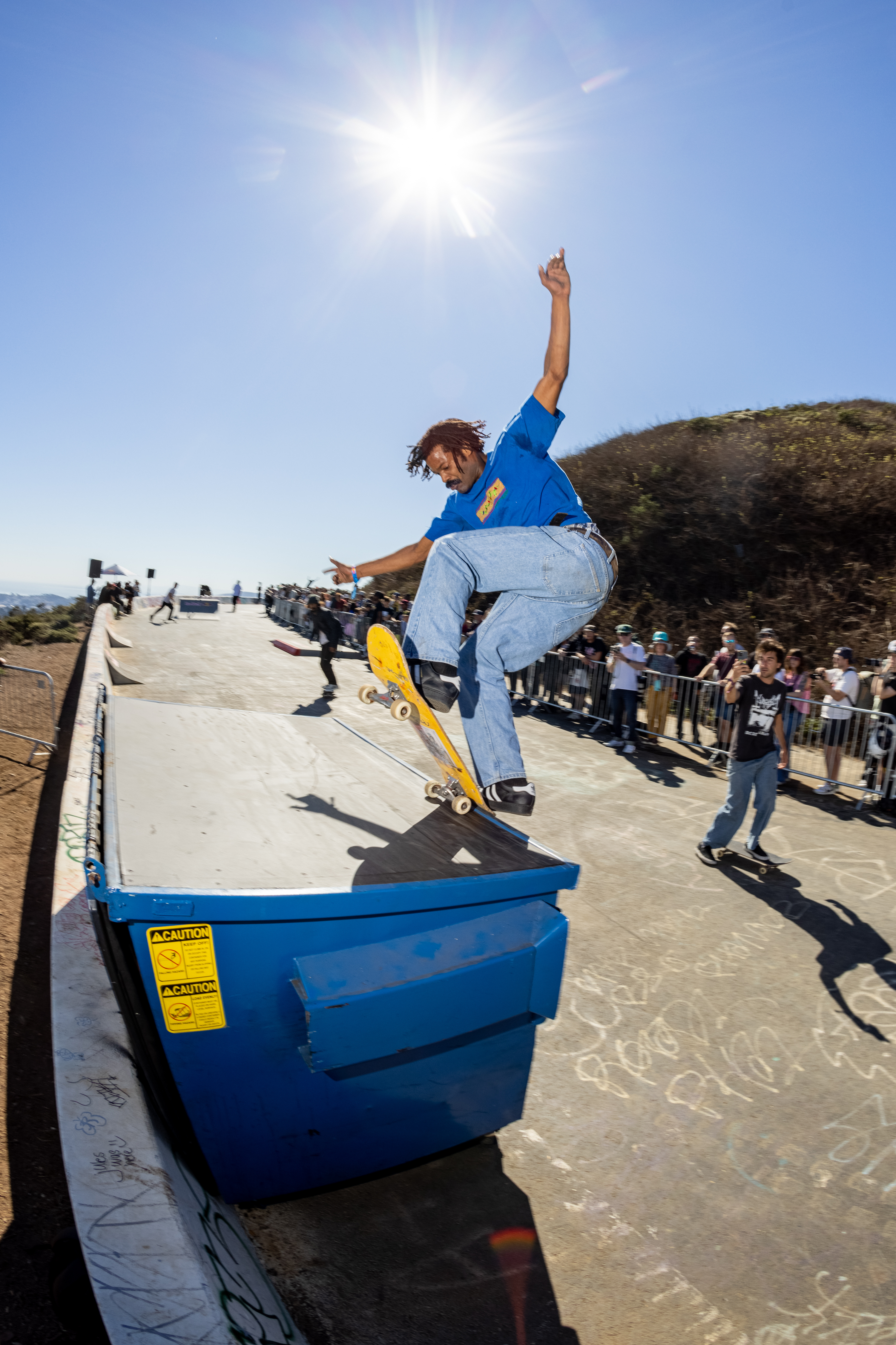 A skateboarder in a blue shirt and jeans performs a trick on a blue dumpster under a bright sun, watched by a crowd. The setting is lively and dynamic.