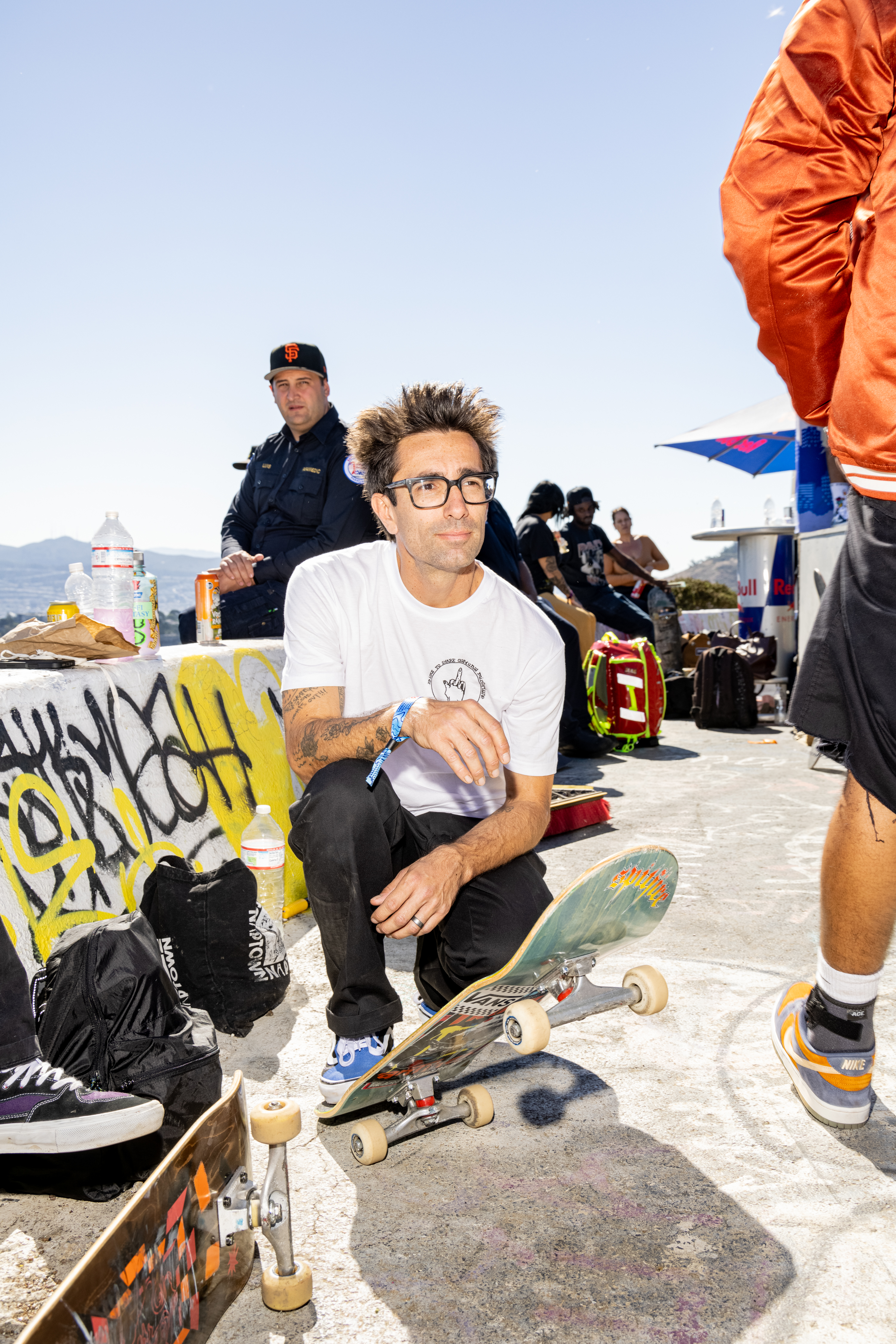 A man in a white T-shirt and glasses crouches with a skateboard, surrounded by graffiti and people. A police officer is in the background.