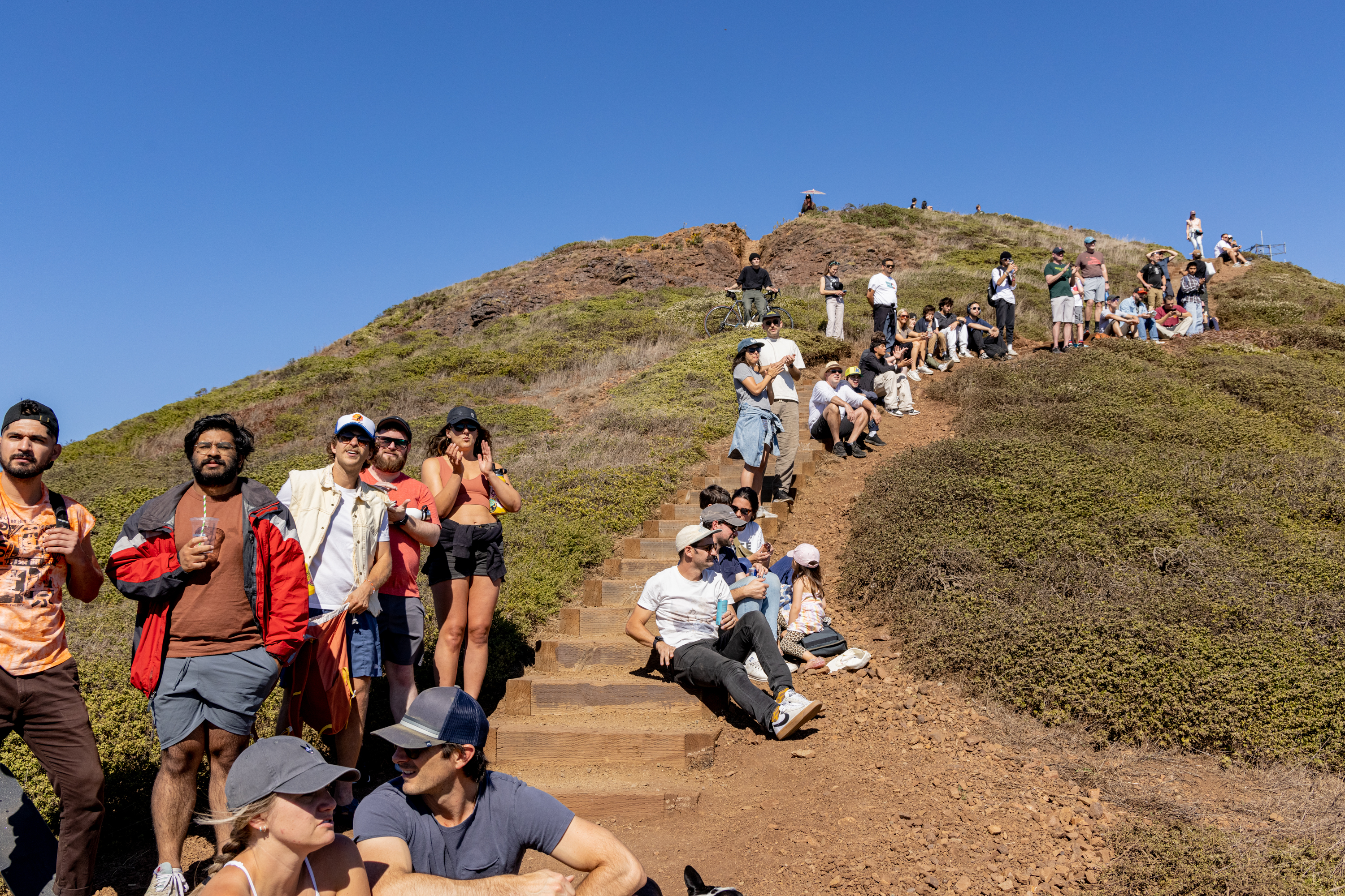 A group of people is gathered on a steep hillside under a clear blue sky, sitting or standing along stone steps, surrounded by brown and green vegetation.